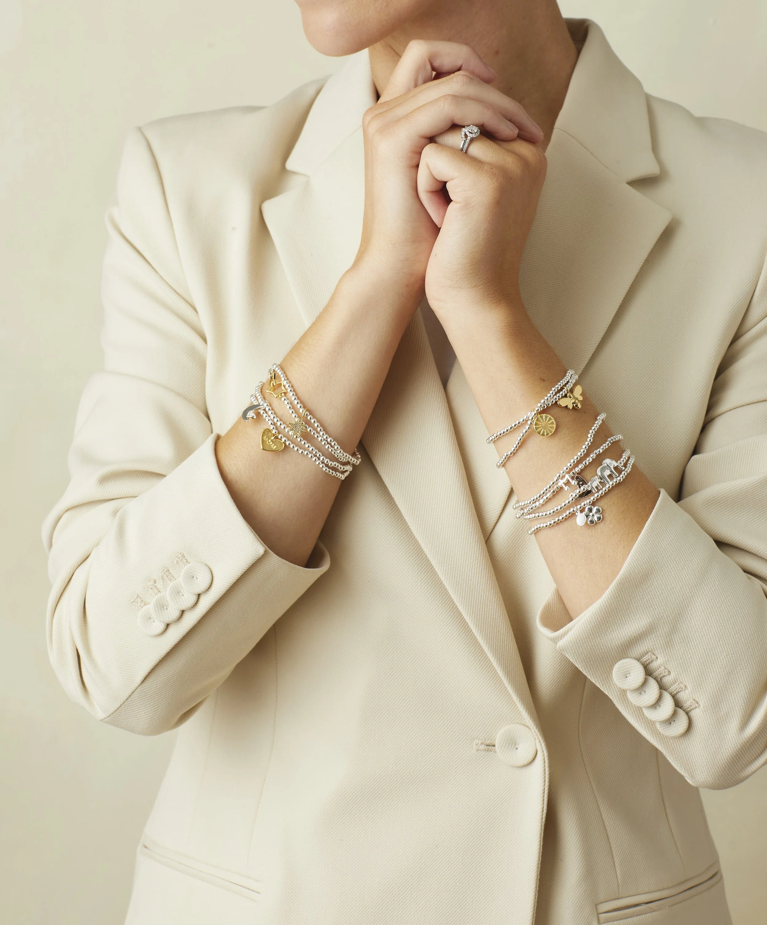 Woman wearing a cream-colored blazer and several silver and gold charm bracelets, with her hands clasped near her chin.