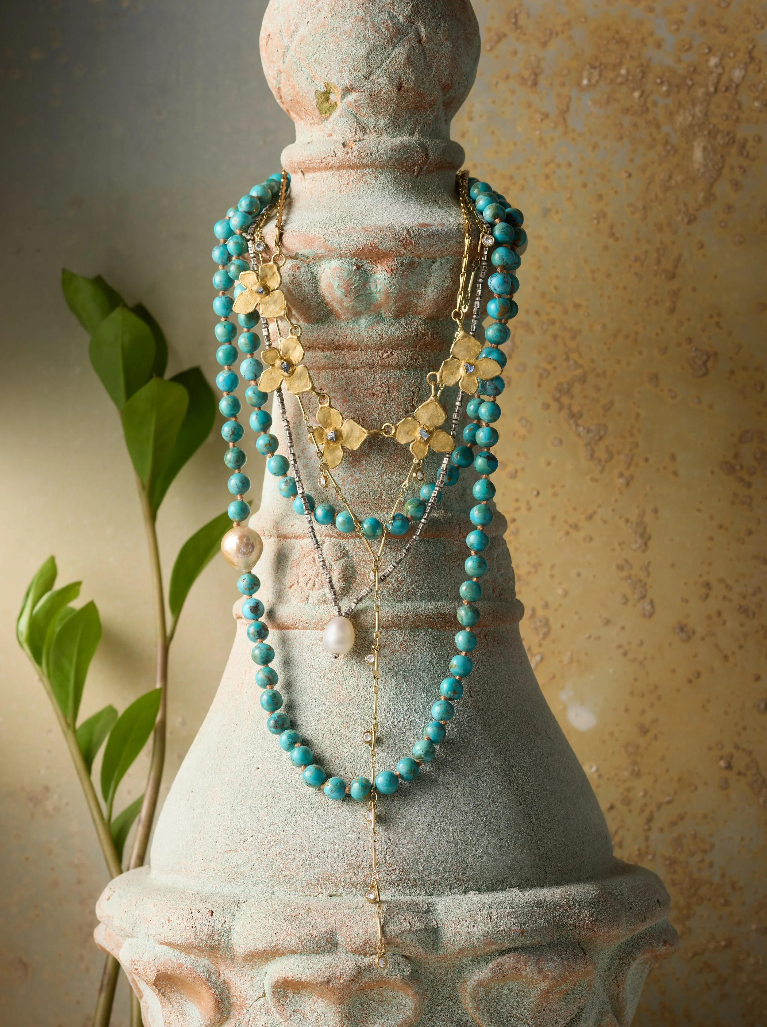 Multiple layered necklaces with turquoise beads, gold flower charms, and pearl pendants draped over a rustic stone bust, with a green leafy plant in the background and a textured beige wall.