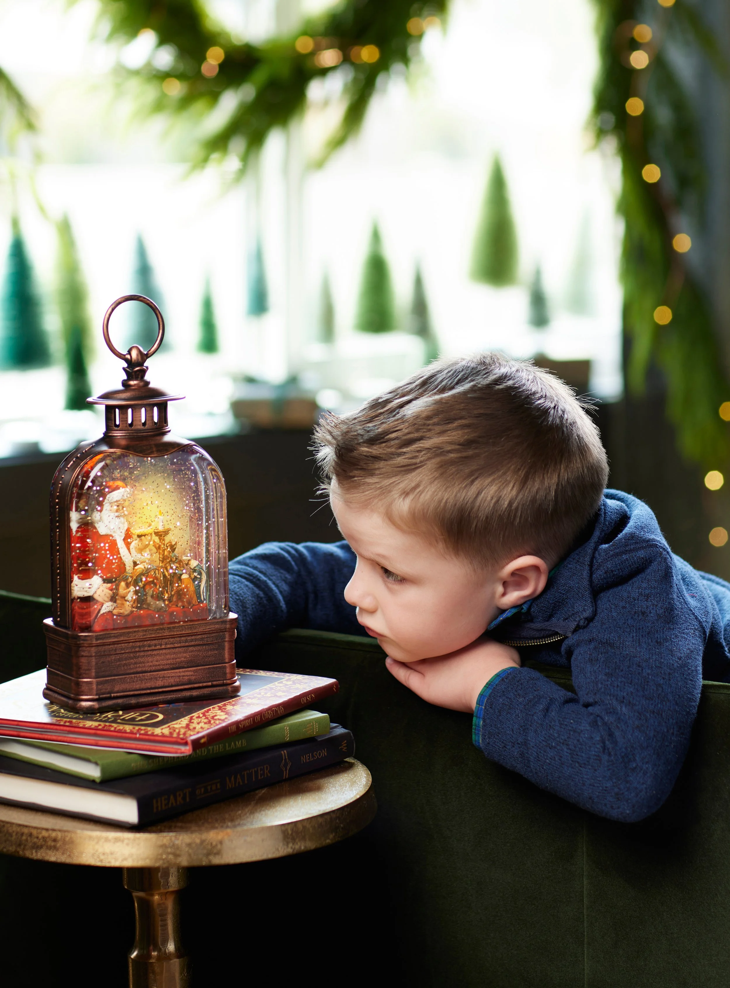 A young boy with short brown hair leaning on a dark green armchair, gazing at a small decorative snow globe with Christmas figures inside, placed on a stack of books on a small round gold-colored table.