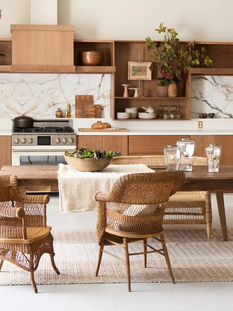 Modern kitchen and dining area with wood cabinets, marble backsplash, woven chairs, dining table with glass jars, and a large bowl of salad.