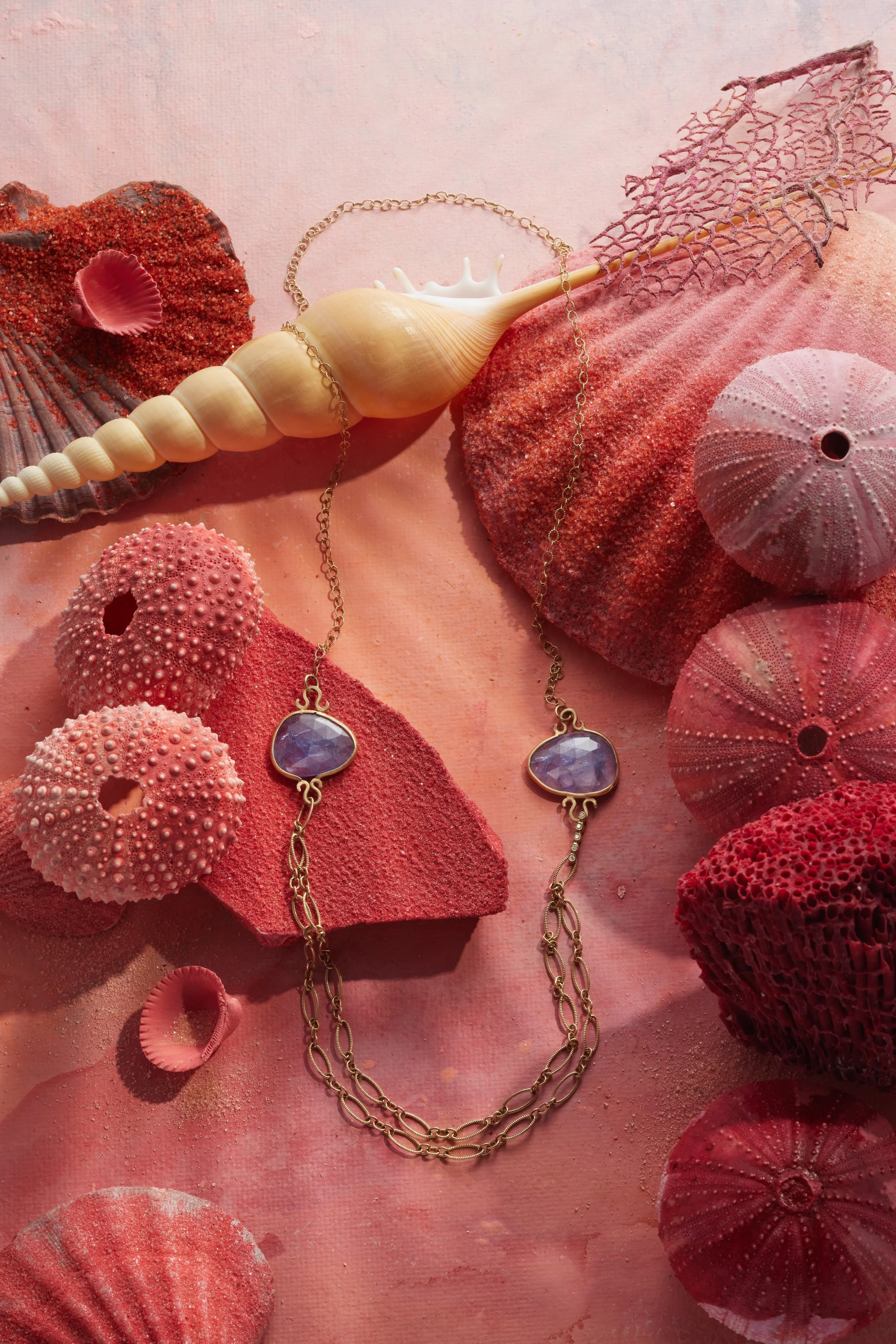 A fall seashell necklace resting on pink sand, surrounded by coral, sea sponges, and shells in pink and red shades.