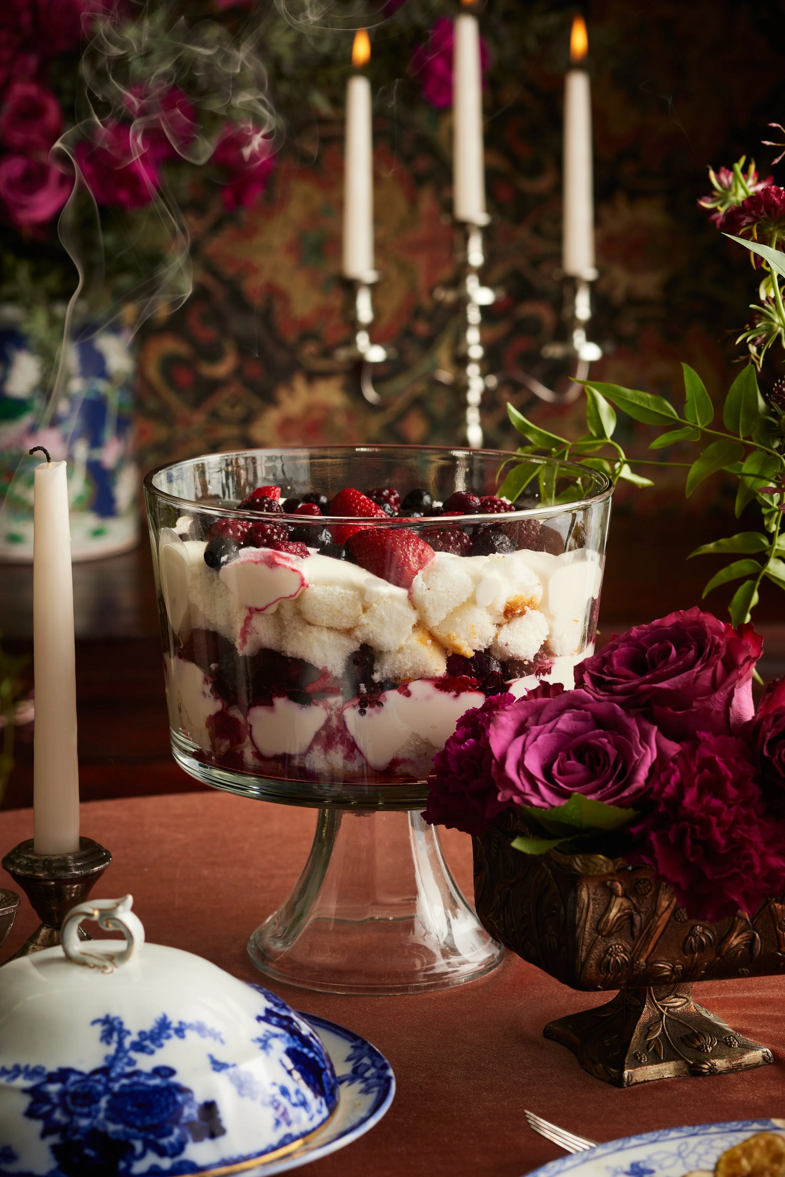 A dessert trifle with mixed berries and whipped cream in a glass bowl on a table decorated with flowers and candles.