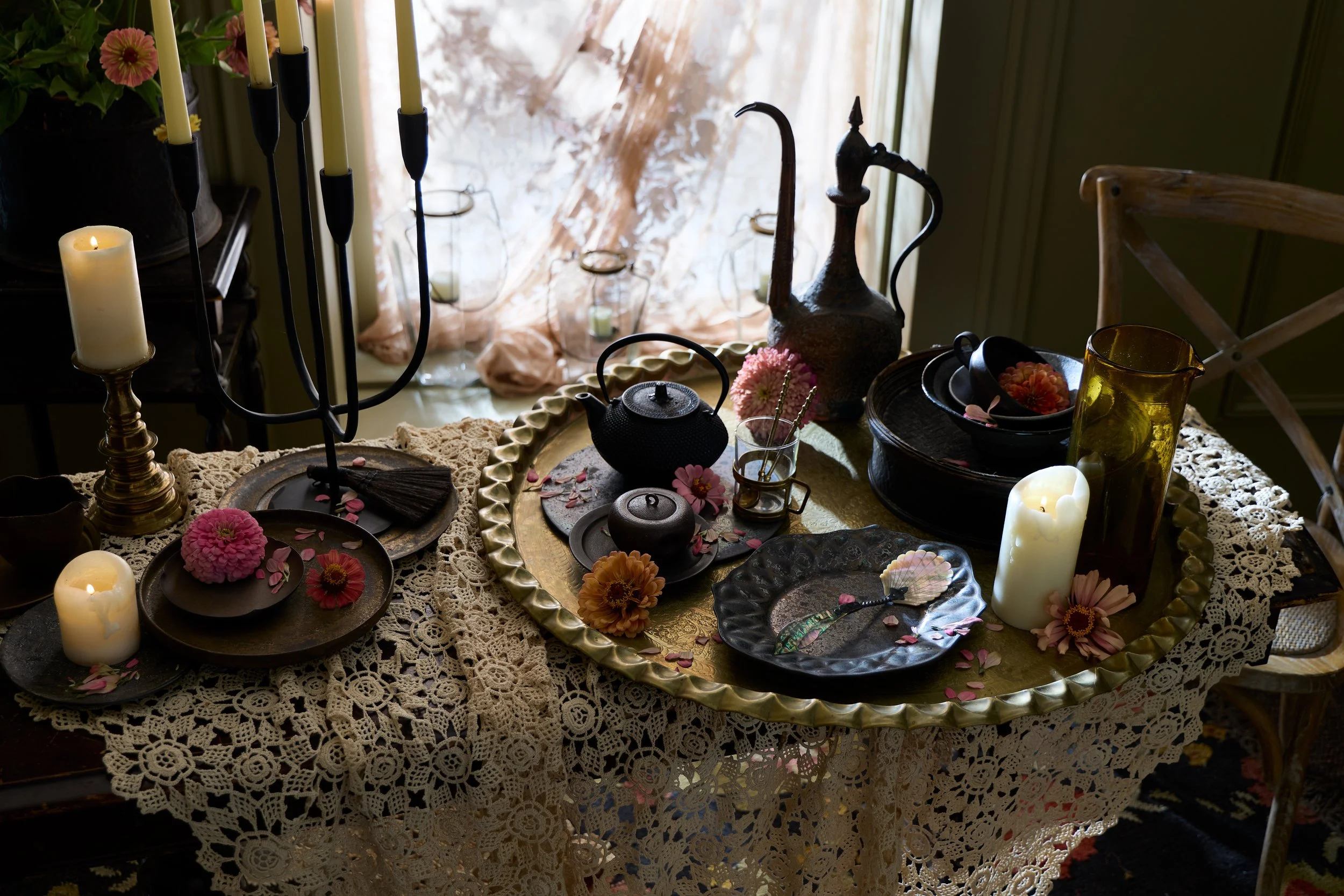 A vintage dining table set with lace tablecloth, decorative plates, candles, flowers, and teapots, in a cozy, softly lit room by a window.