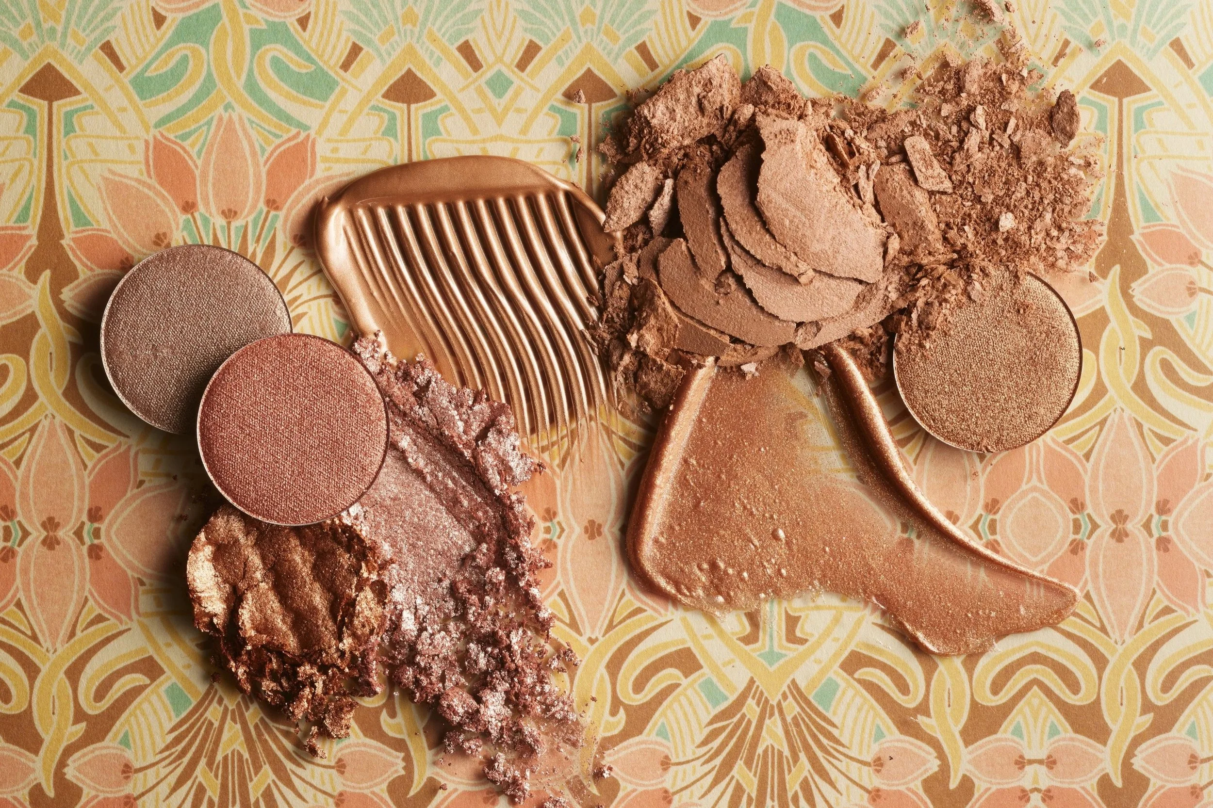 Various shades of pink, bronze, and brown makeup products including eyeshadows, blushes, and highlighters arranged on patterned background.