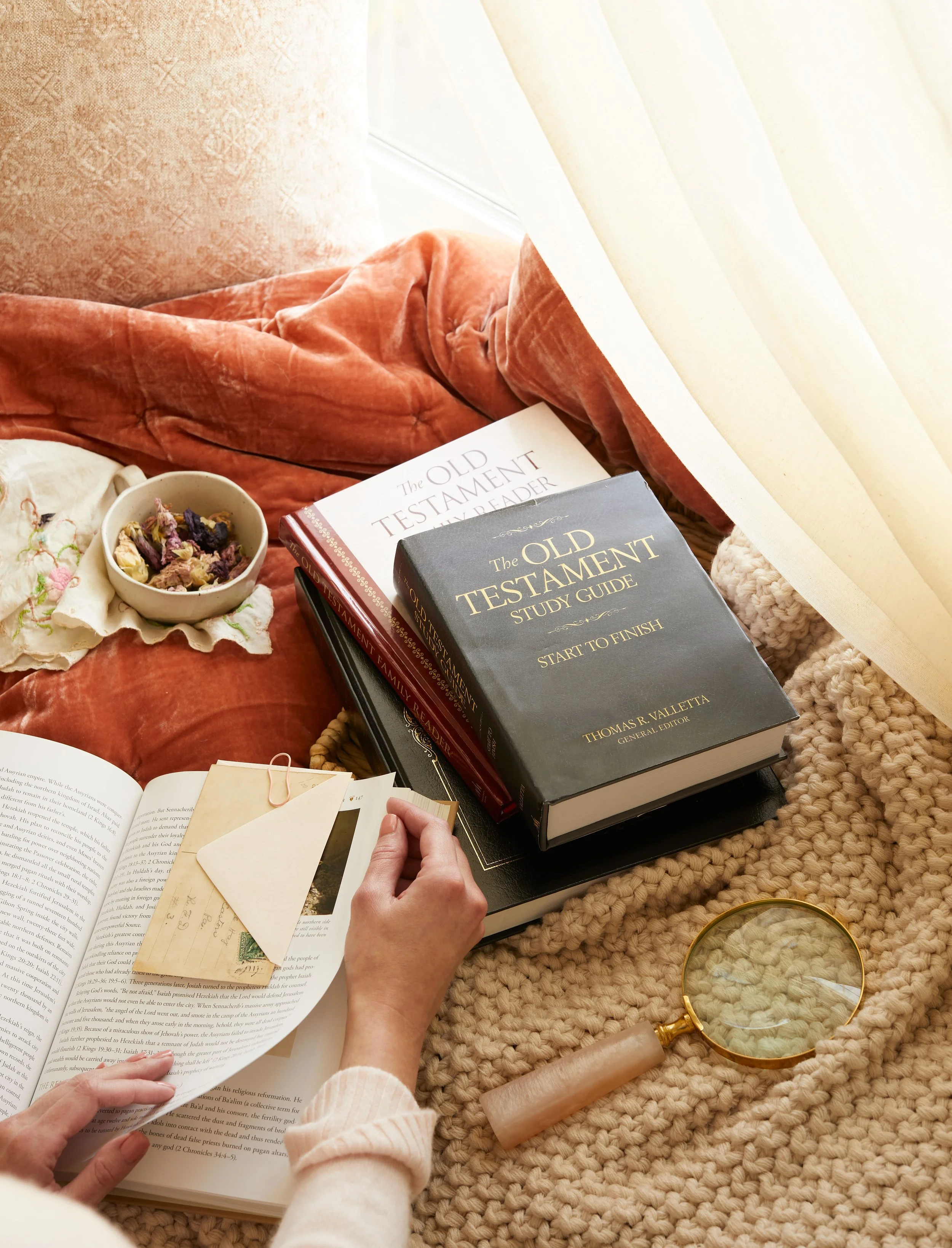 Open Bible with a person's hand holding a page marker, a set of stacked books including 'The Old Testament Study Guide,' a bowl with dried herbs or potpourri, a magnifying glass, and a cozy, plush blanket on a cushioned surface near a window with curtains.