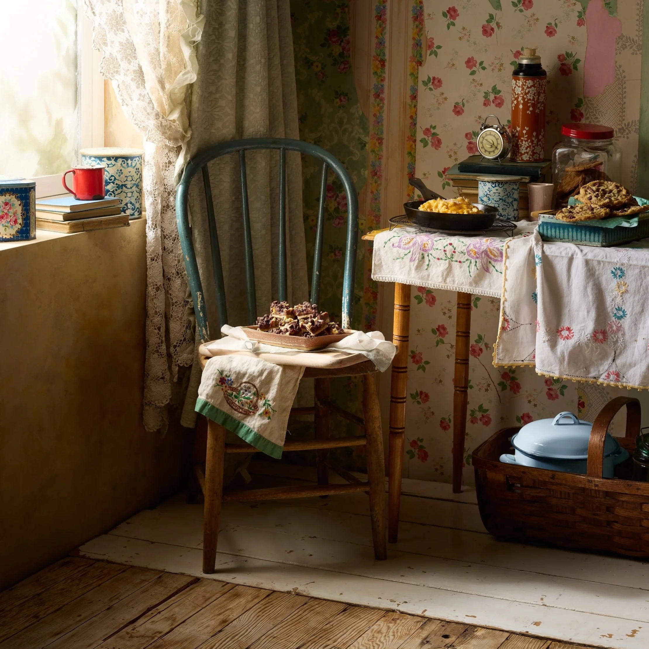 A cozy kitchen corner with a vintage wooden chair with a cloth on it, a small side table with a tablecloth, and various kitchen items including a cast iron skillet, a thermos, a stack of books, and a jar of cookies. There are lace curtains by a window with sunlight filtering in.