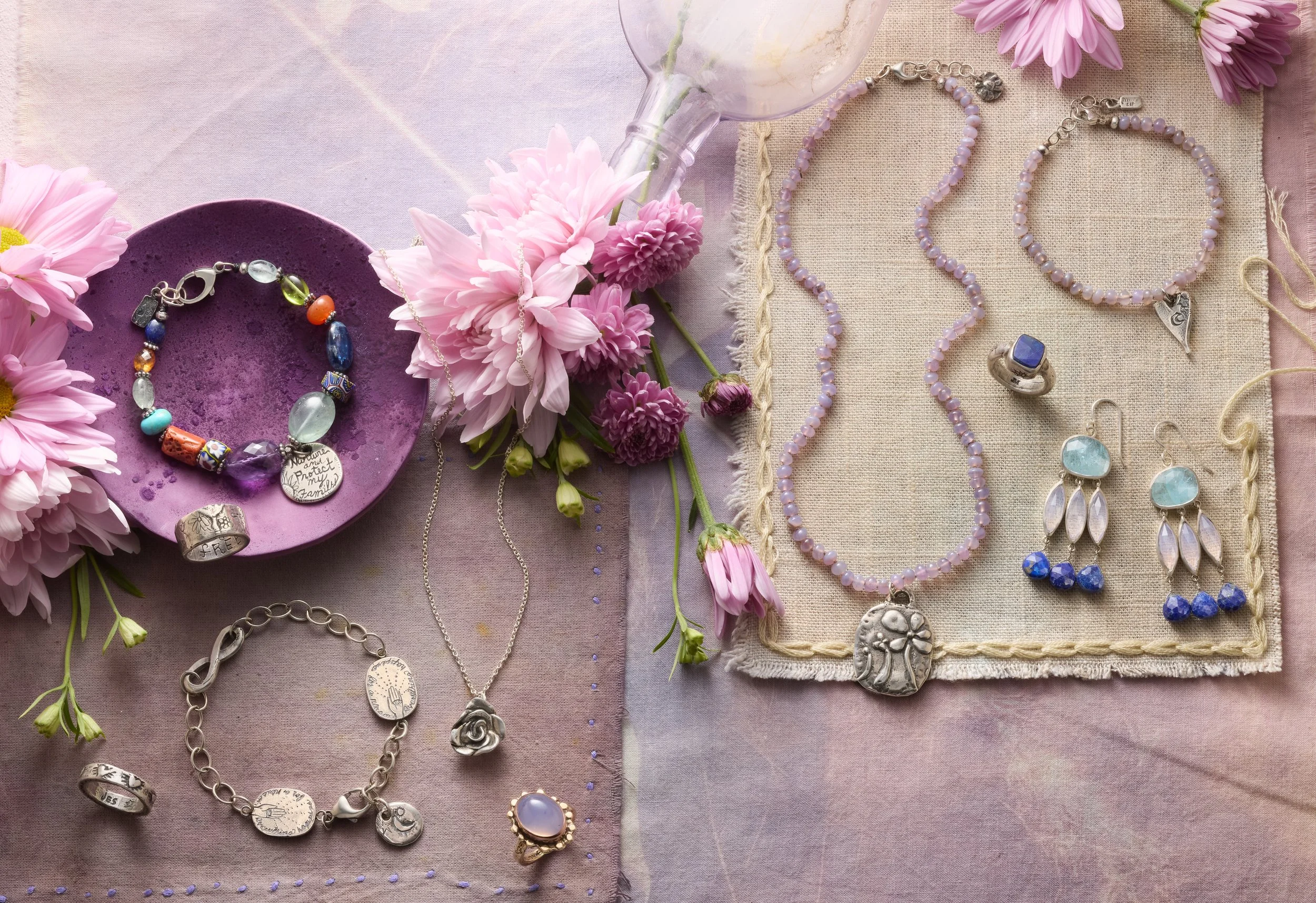 Jewelry display with pink flowers, necklaces, earrings, rings, and a bracelet on a pastel fabric surface.