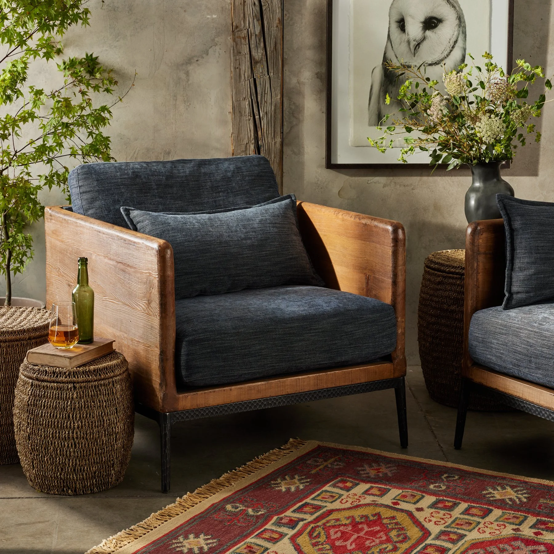 Living room corner with two modern armchairs with wooden armrests and dark fabric cushions, a large framed owl portrait on the textured wall, a green potted tree, a small woven side table with a drink and a book, a large vase with flowers, and a decorative rug.