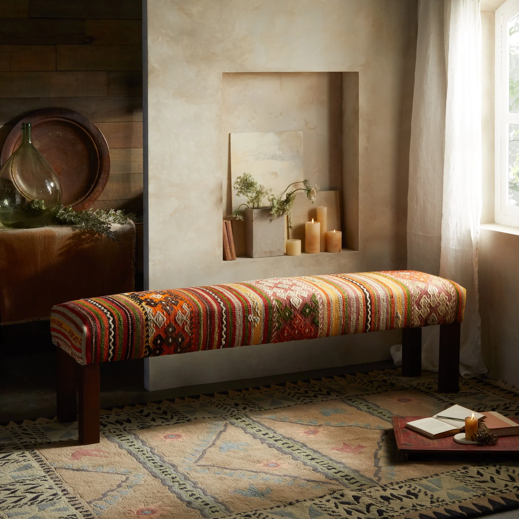 A cozy room corner with a colorful, patterned upholstered bench against a textured beige wall, illuminated by natural light from a window, with a decorative tray containing a candle and book on the floor, and a display of candles and plants on a recessed wall shelf.
