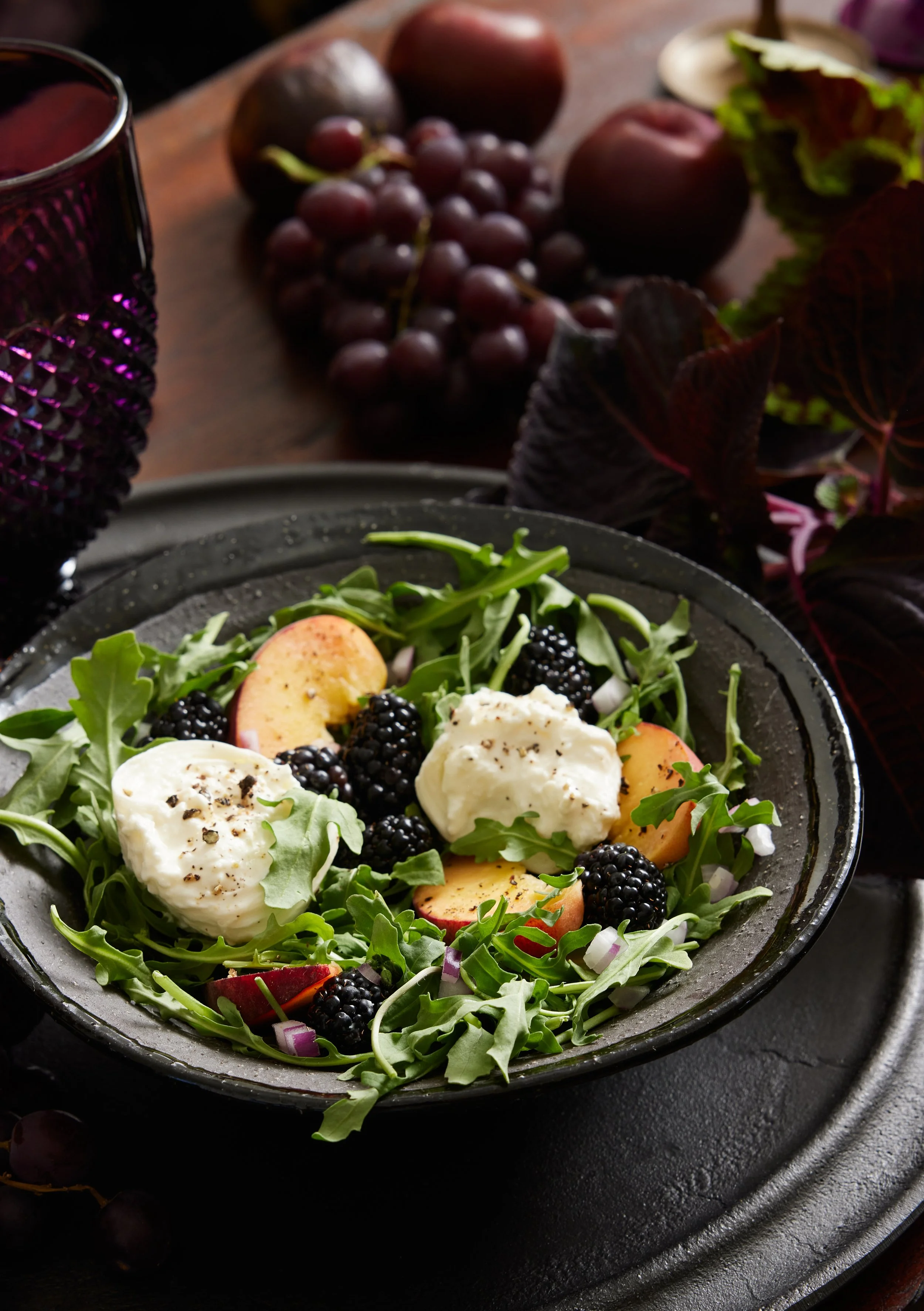Salad with arugula, blackberries, apple slices, and cheese, accompanied by grapes and apples on a wooden table.