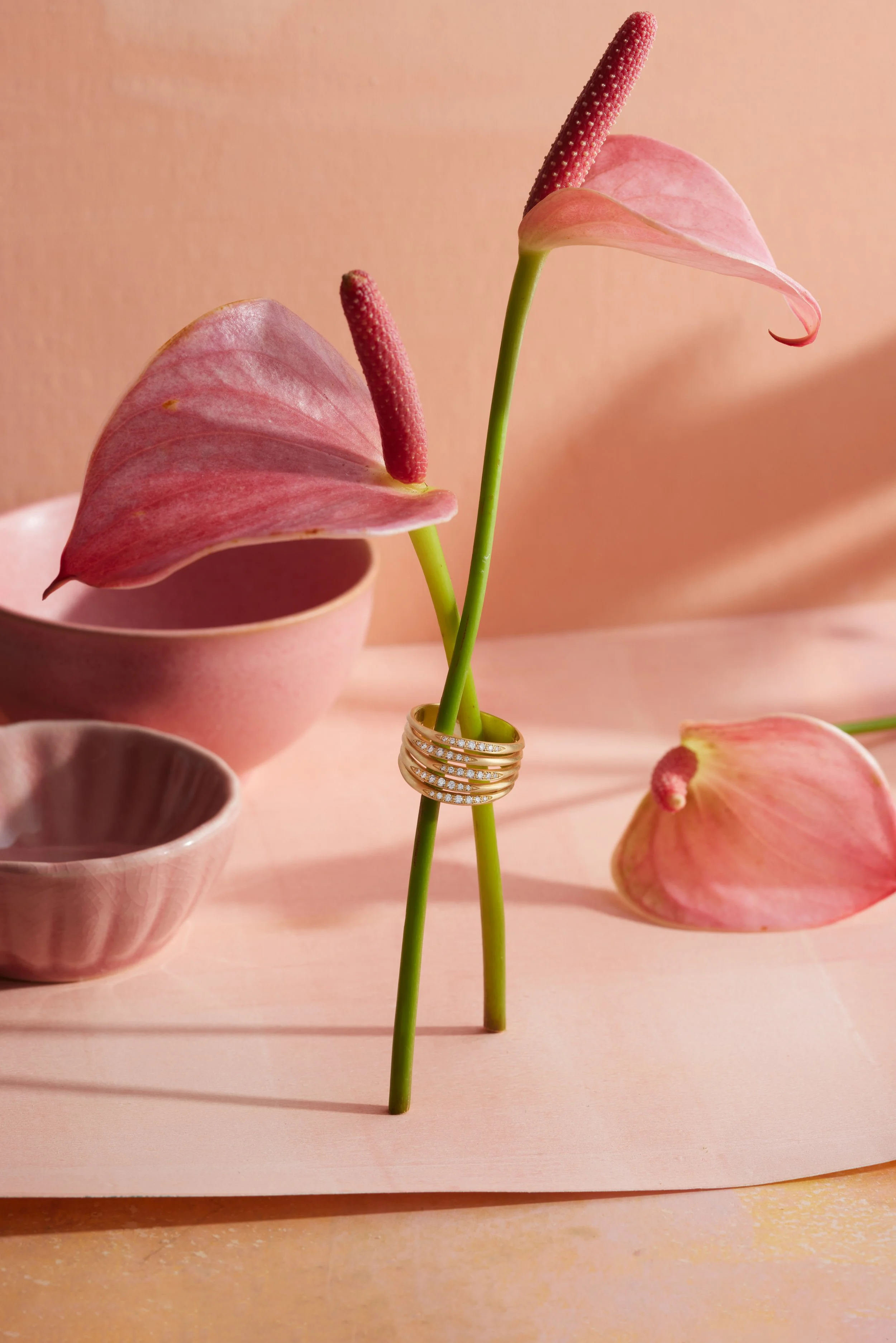 Pink anthurium flowers with green stems, one of which is adorned with a diamond ring, set against pink ceramic bowls and a pink background.