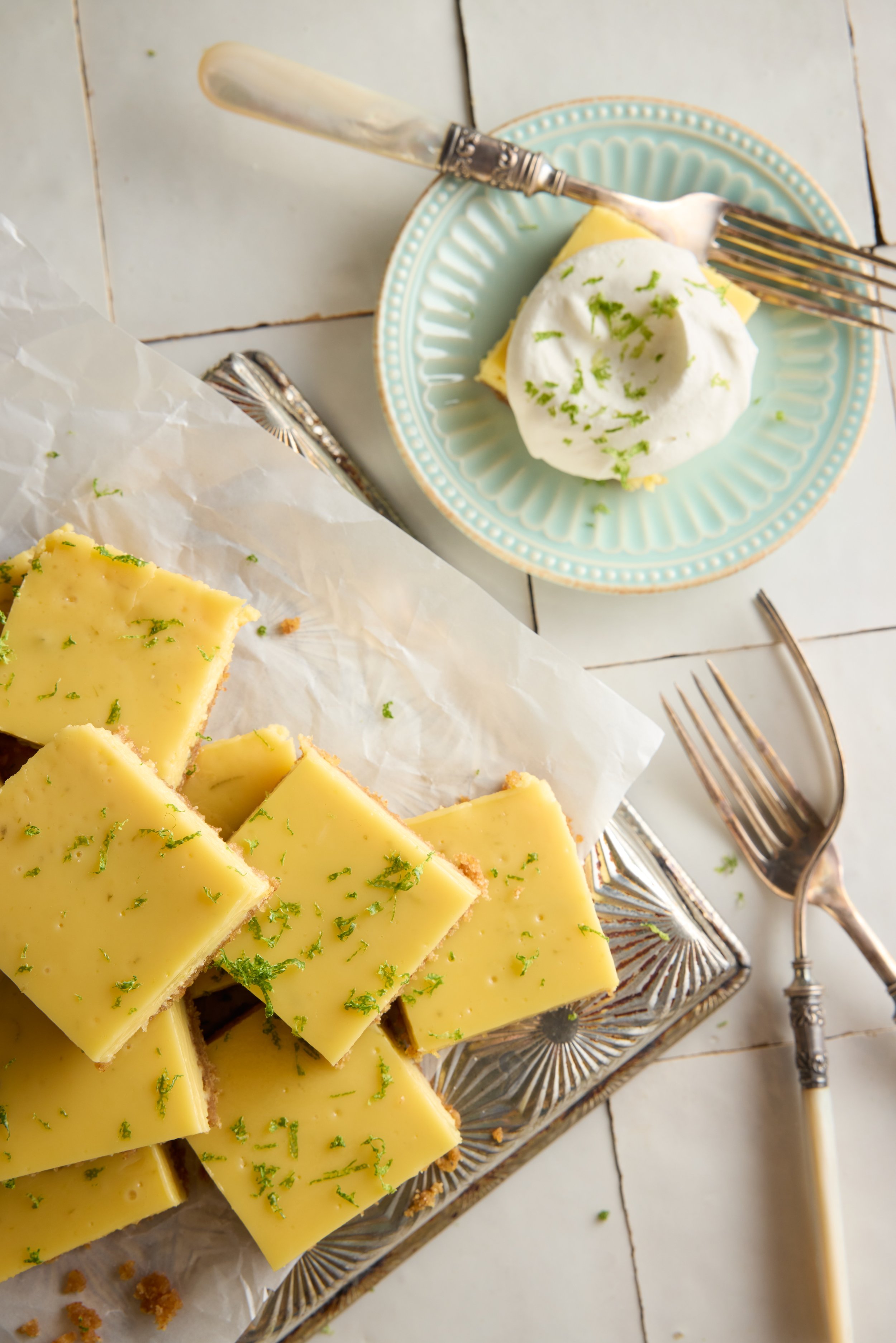 Lemon bars topped with lime zest on a glass tray, with a plate of whipped cream garnished with lime zest on a light blue plate, set on a white tiled surface with vintage forks.