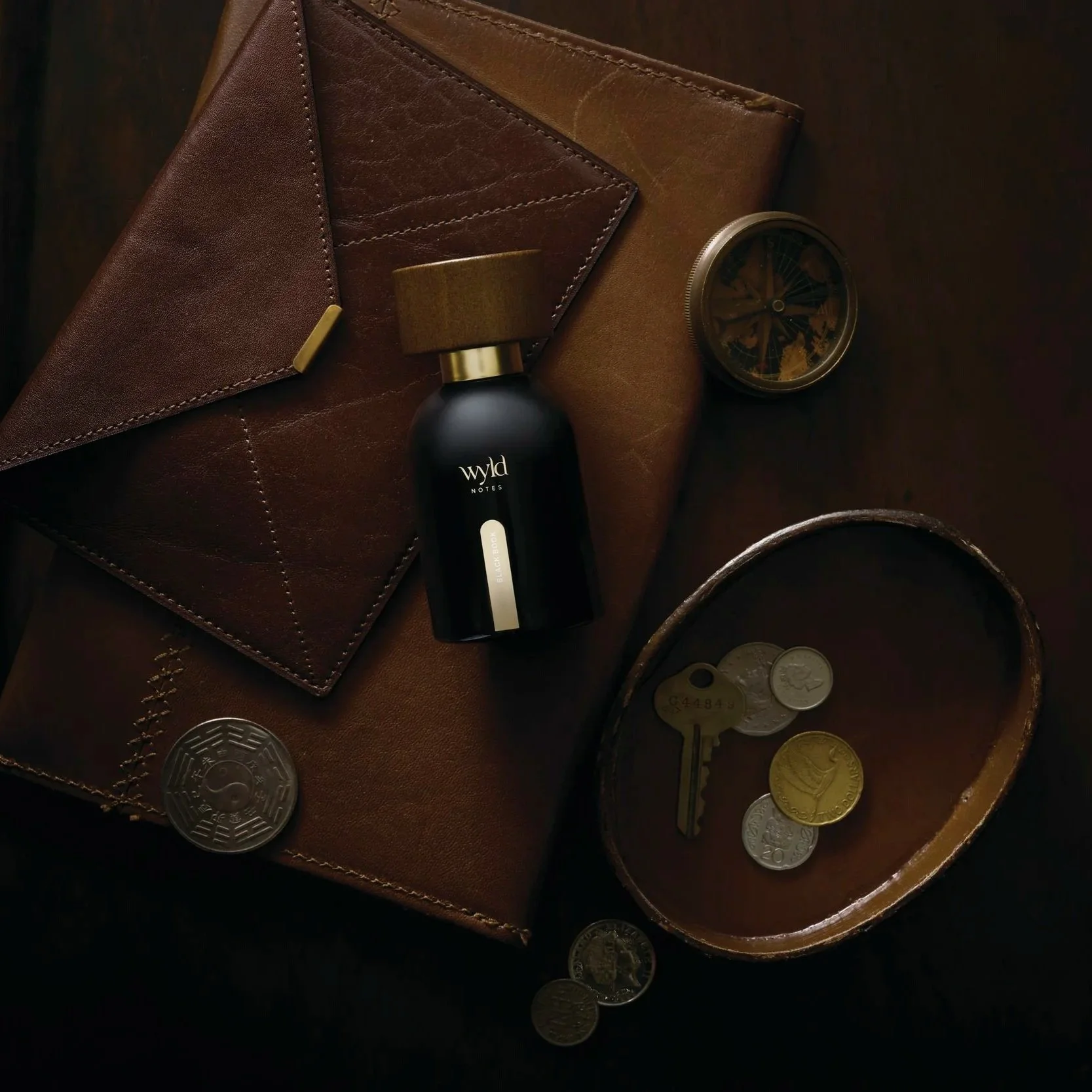 A leather wallet, a perfume bottle, a small pocket compass, and a bowl of coins on a dark surface.