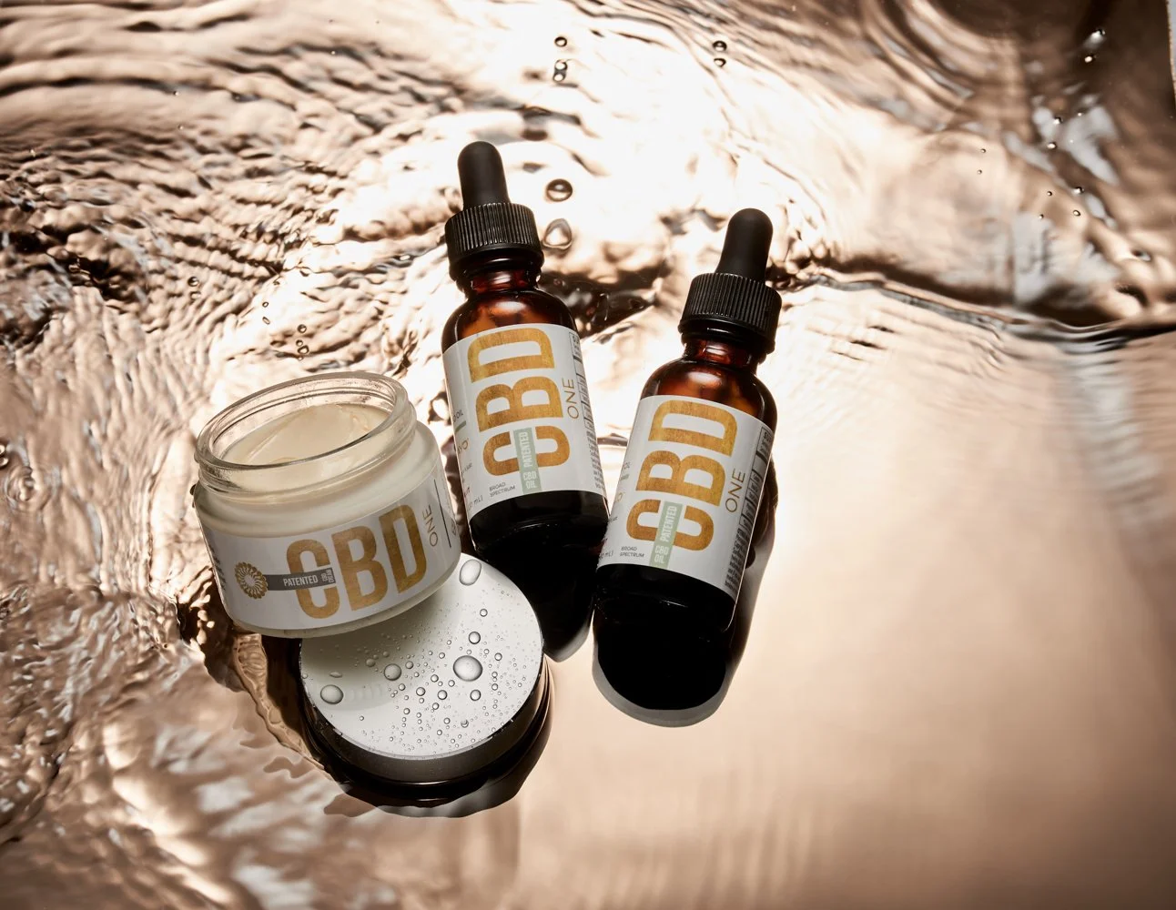 CBD products, including a jar, two dropper bottles, and a circular mirror, on a reflective, textured surface with ripples and bubbles.