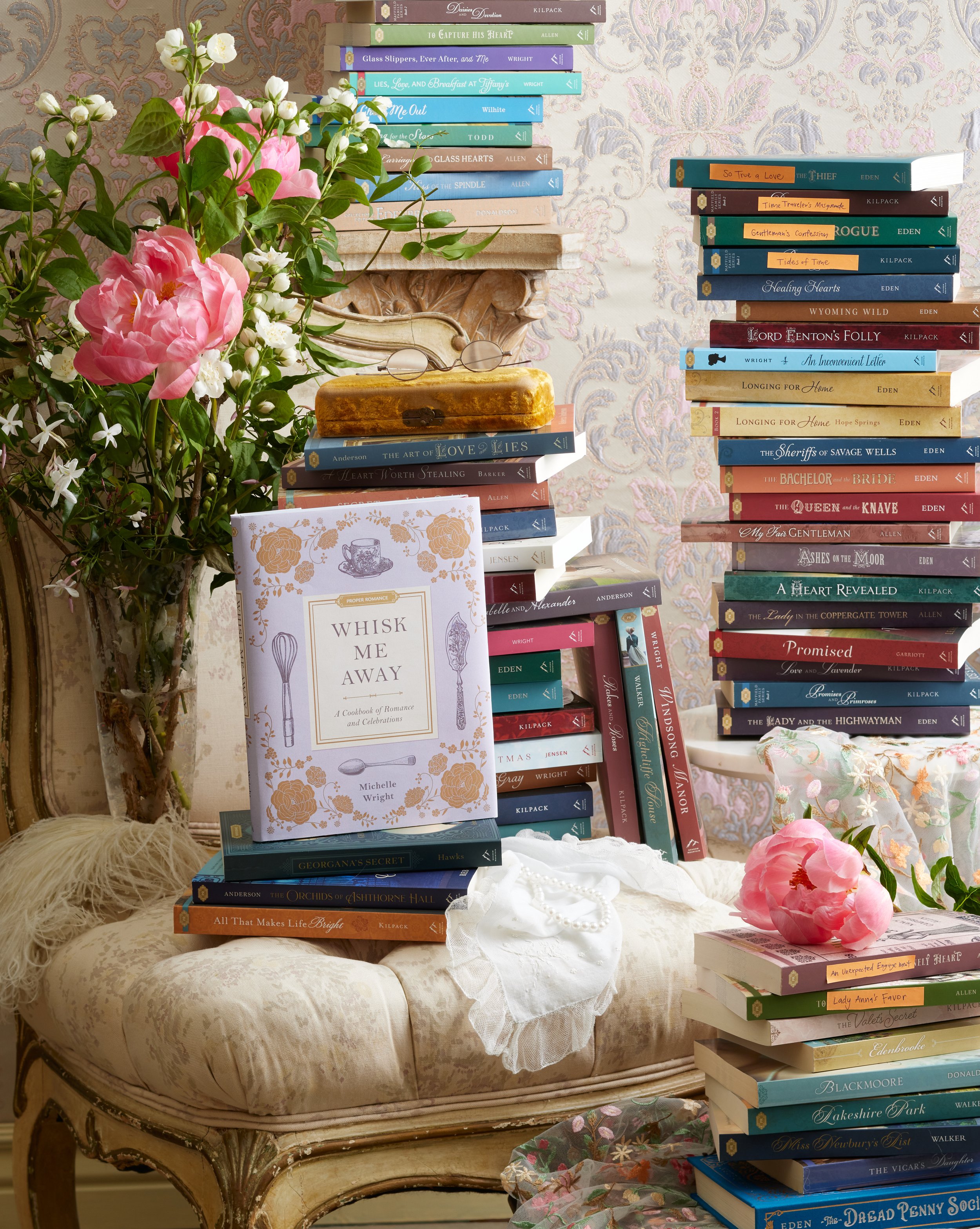A vintage-style sitting area with a floral arrangement, stacks of colorful books, and a pink peony flower on a cream-colored ottoman.