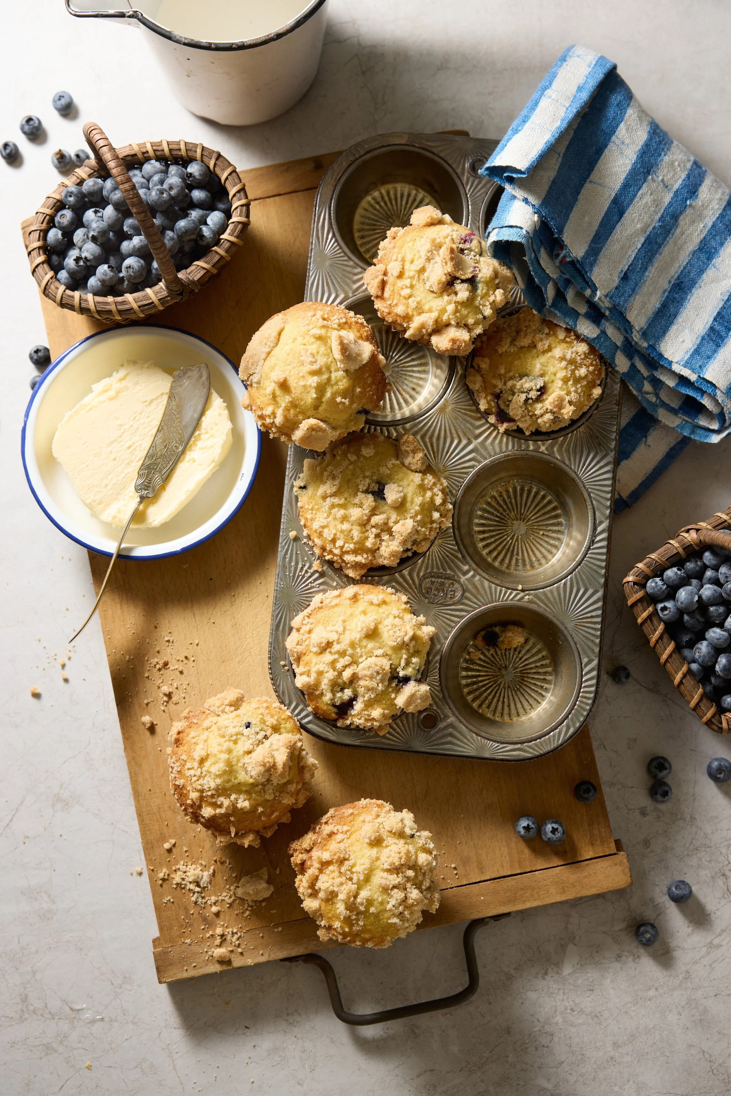 Freshly baked blueberry muffins on a baking tray, with a bowl of butter, a basket of blueberries, and a cup of milk nearby on a wooden cutting board, with a blue and white striped cloth.