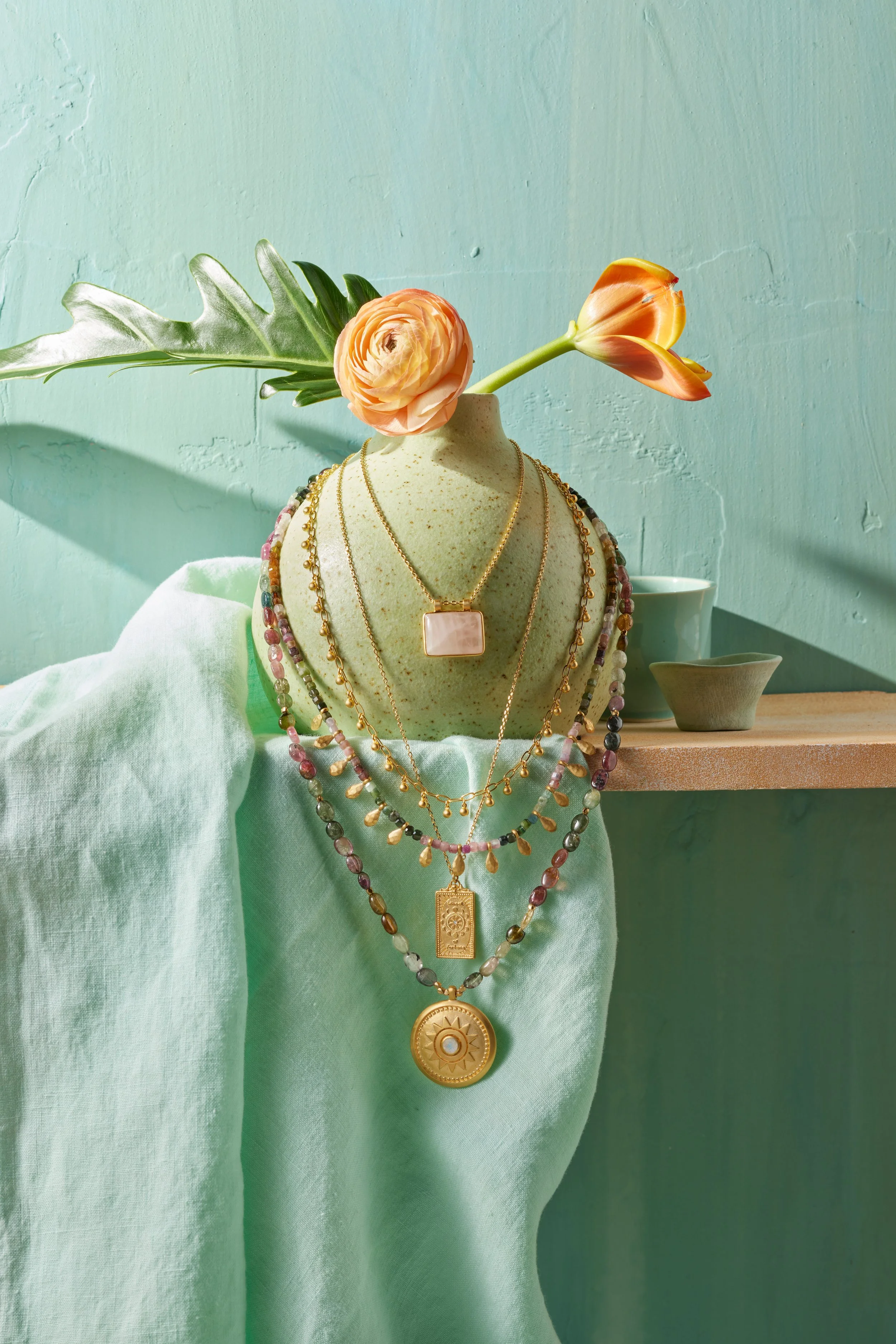 Display of layered gold and beaded necklaces on a jewelry bust with a light peach stone, a peach ranunculus, and an orange tulip, draped with a light green cloth, against a mint green wall.