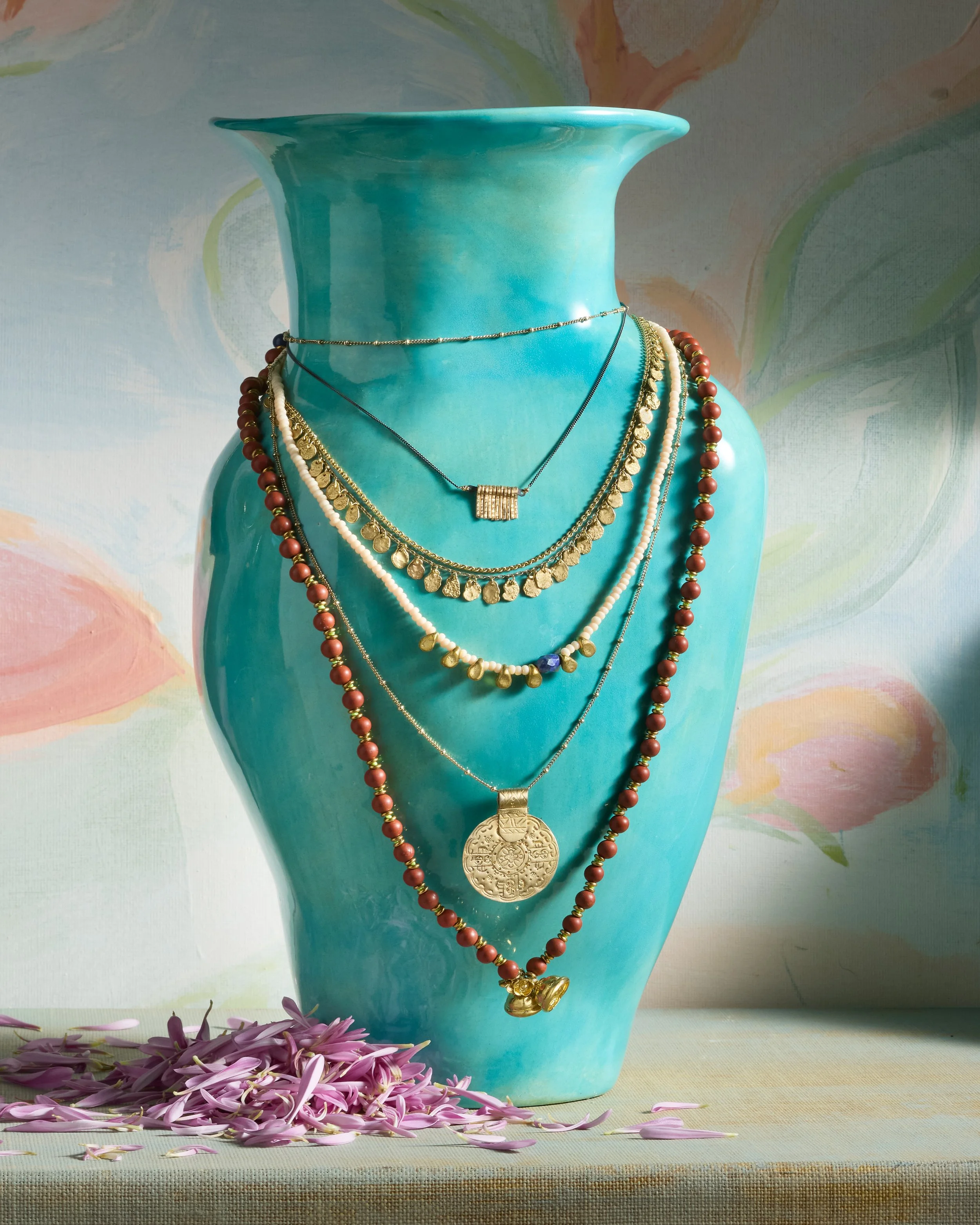 A blue ceramic vase displays multiple layered necklaces of various styles, including gold, pearl, and beaded designs, with pink flower petals scattered at the base.