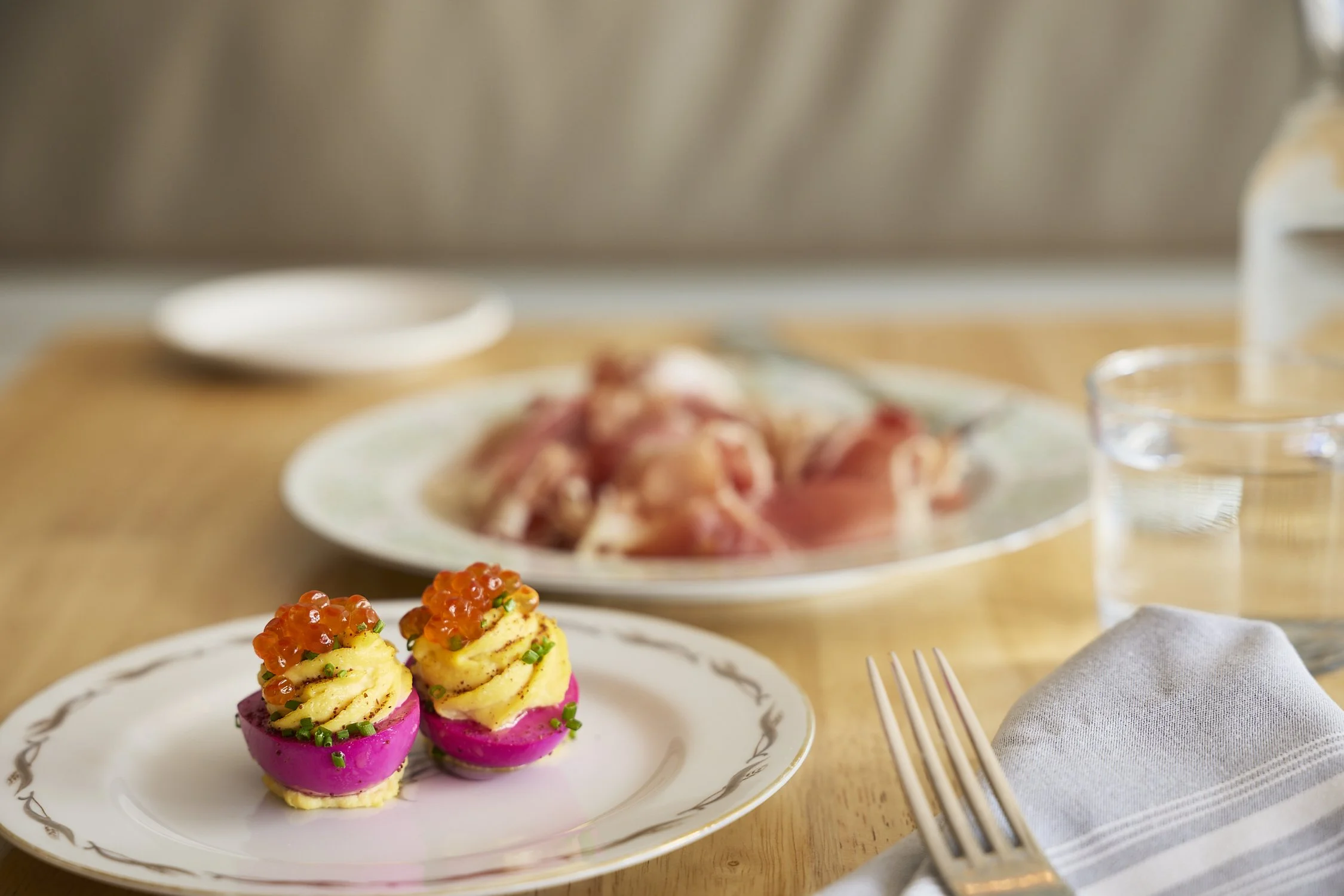 Colorful canapé appetizers with pink base, topped with mashed potatoes, orange fish roe, and green herbs, placed on a white plate with gold rim.