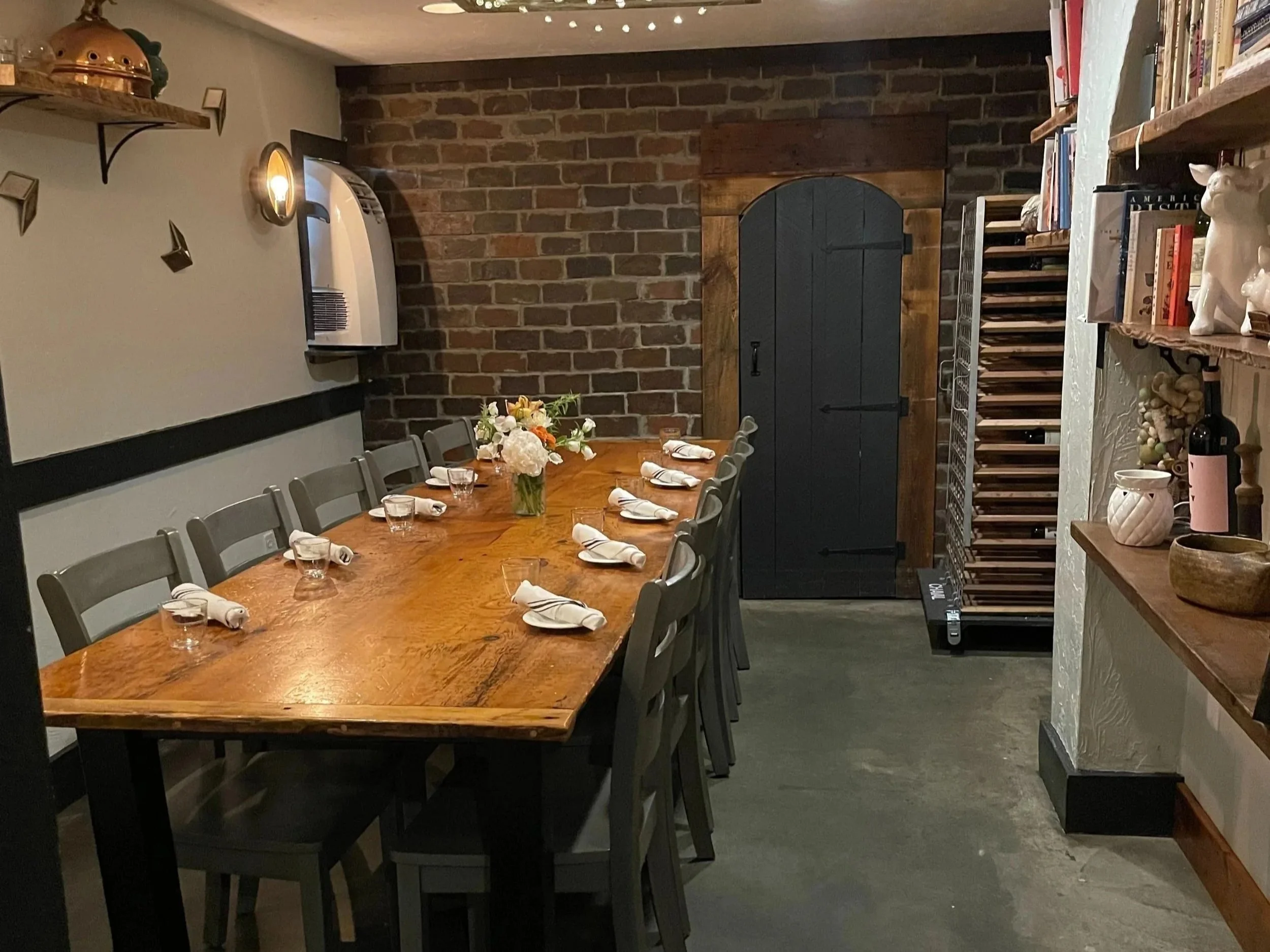 A dining room with a long wooden table set with white napkins, glasses, and a floral centerpiece. There are grey chairs around the table. The room has a brick wall, a wooden door, and shelves with books and decorative items. The lighting is soft, with a wall-mounted sconce and ceiling lights.