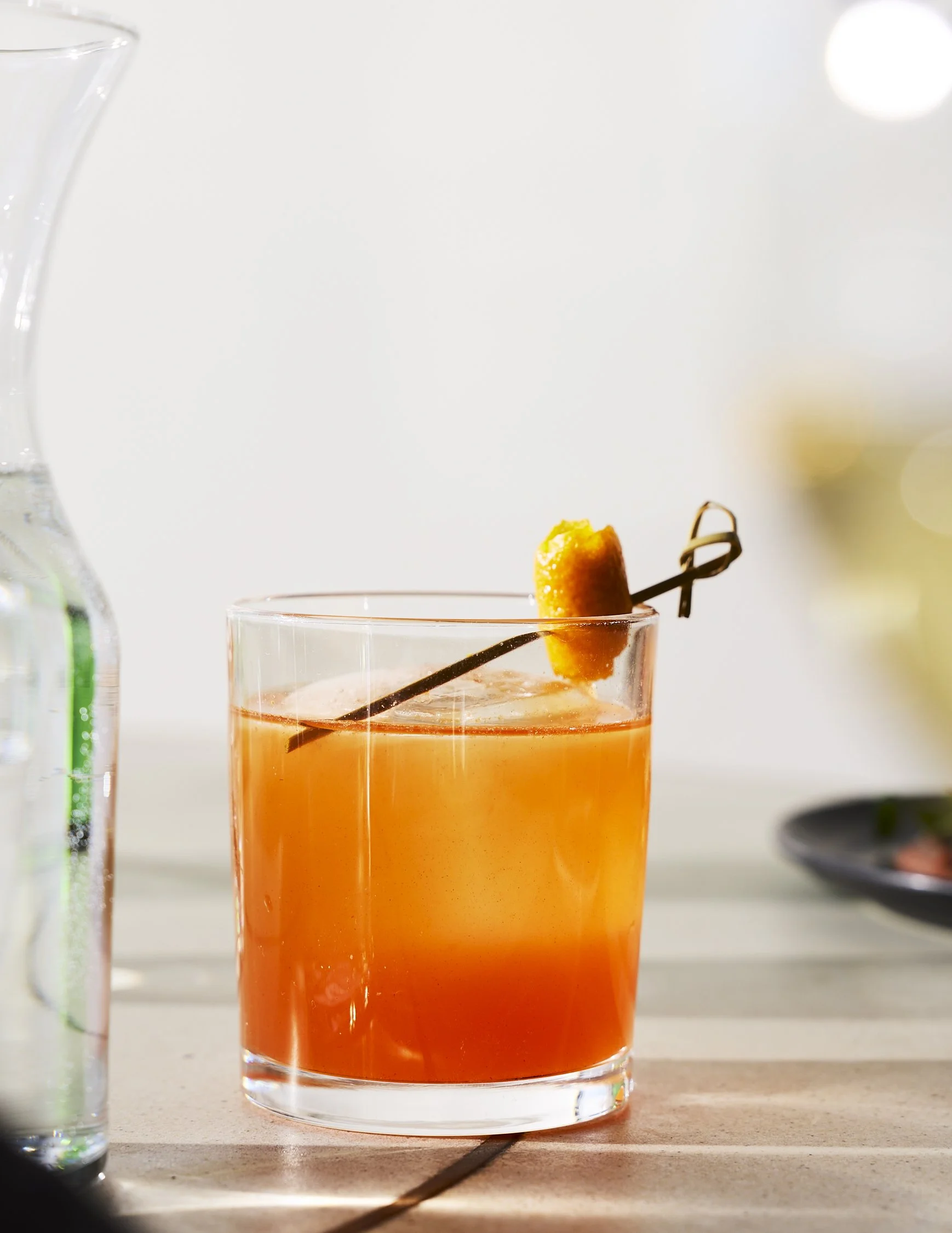 A glass of orange cocktail with a garnish on a white surface, with a blurred background and a partial water bottle visible on the left.