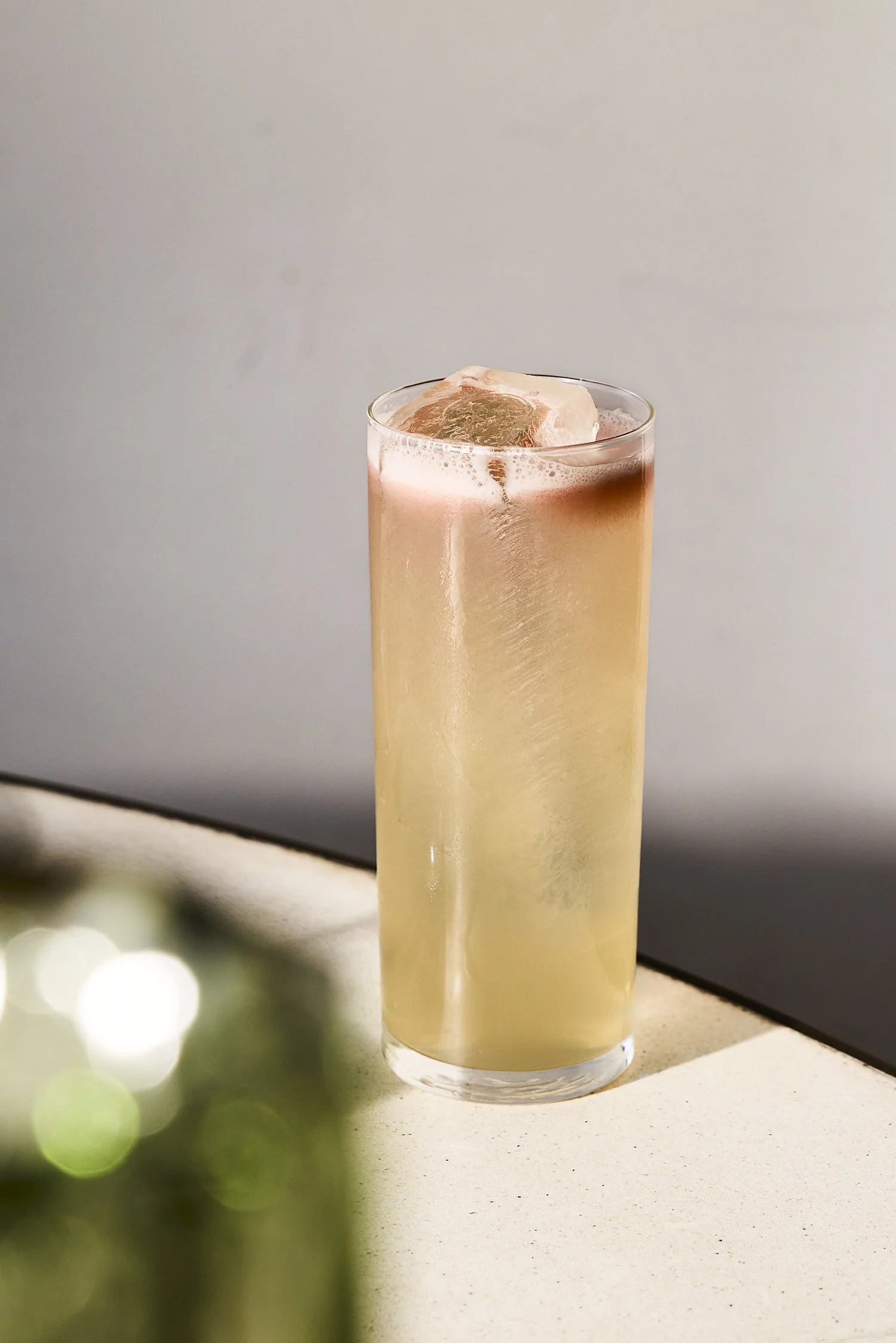 A tall glass filled with a light-colored beverage and ice cube on top.