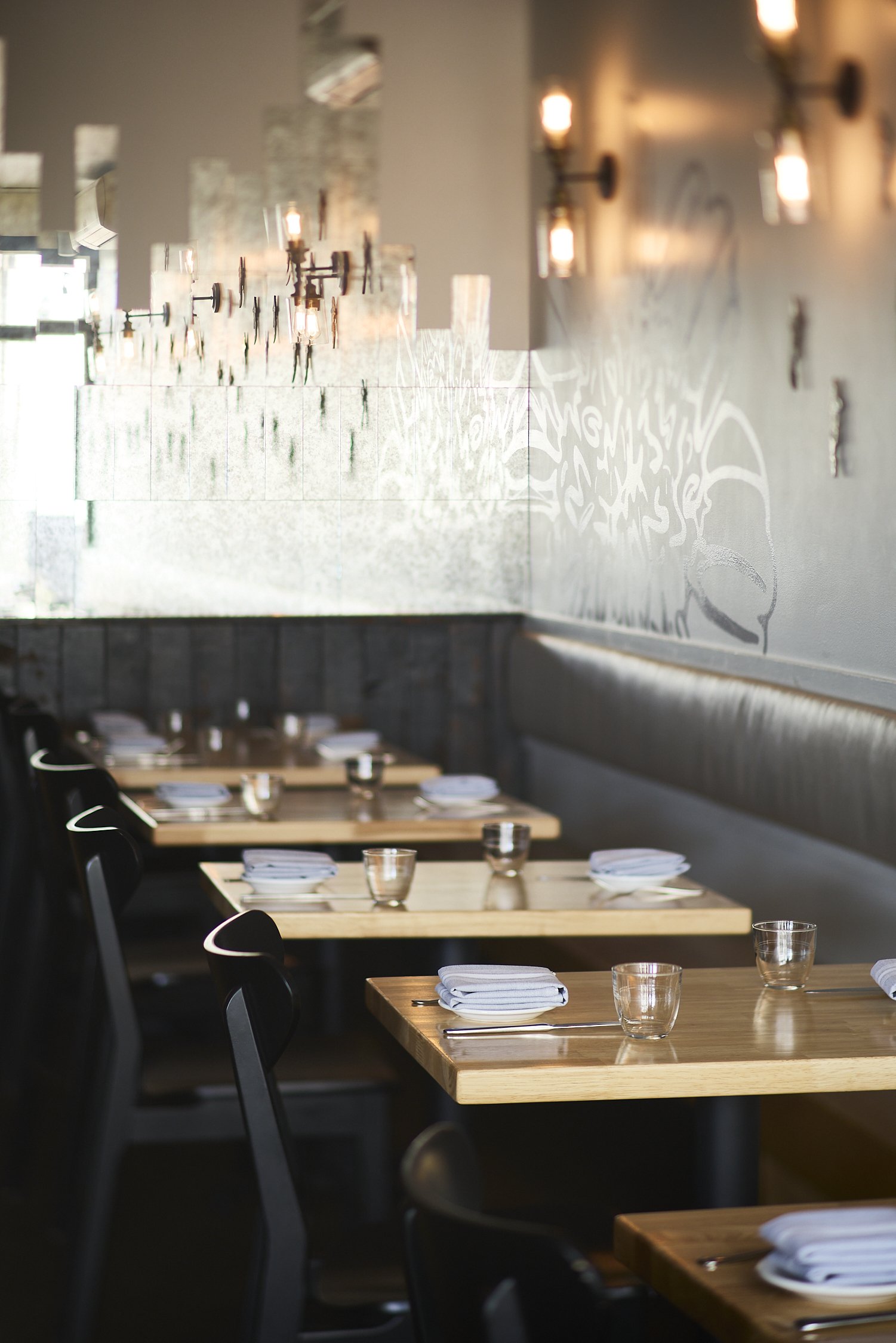 Empty restaurant dining room with wooden tables set with white napkins, glasses, and chairs, decorated with modern wall-mounted lights and artwork.