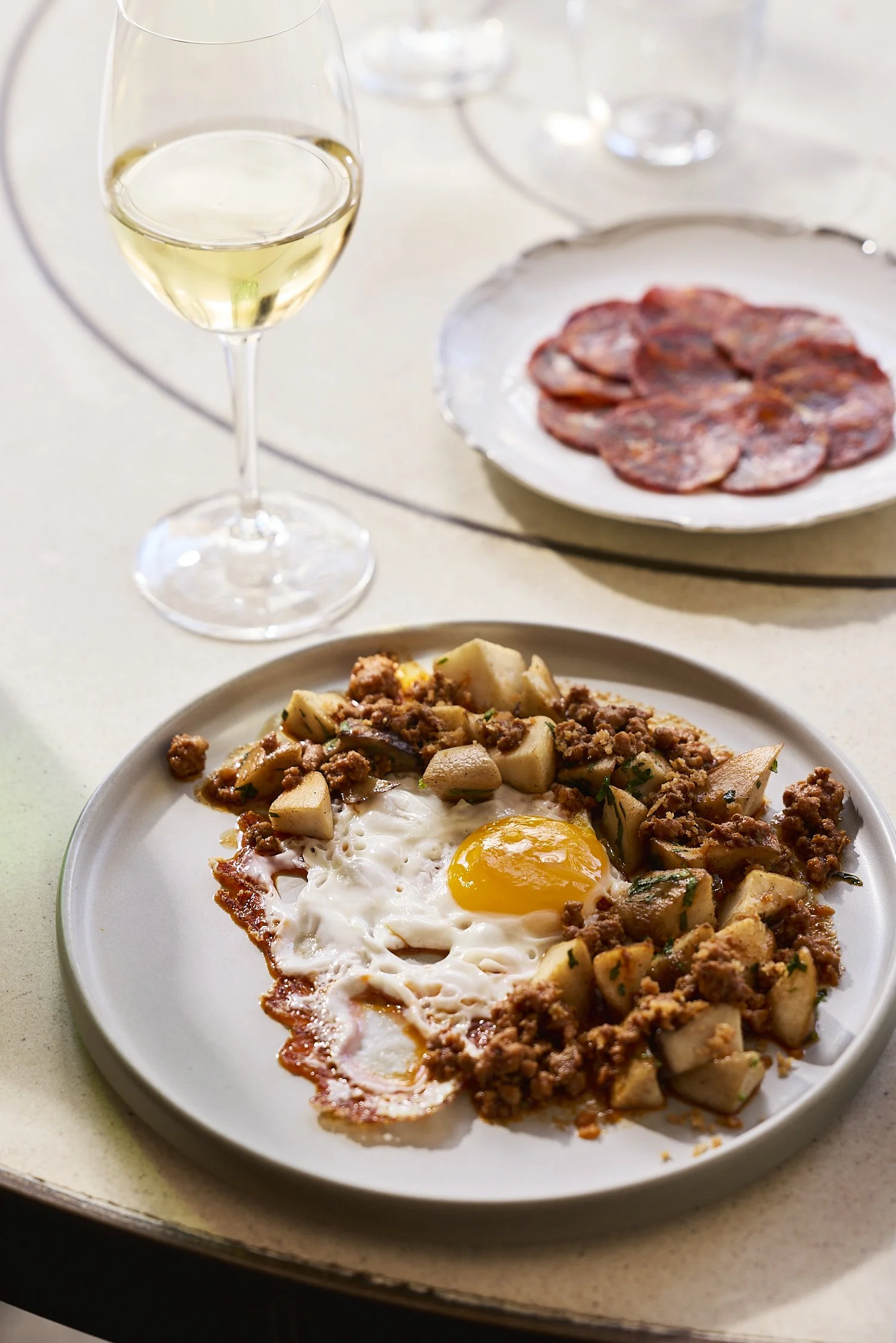 A plate with a fried egg, sausage, and potatoes, in front of a glass of white wine and a plate of sliced cured meat.
