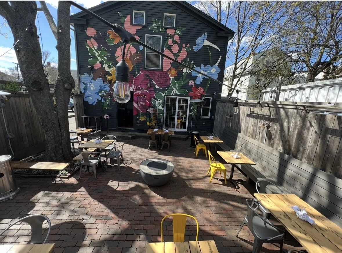 An outdoor patio with wooden tables and chairs, string lights, trees, and a mural of colorful flowers and leaves on the dark-colored building wall in the background.