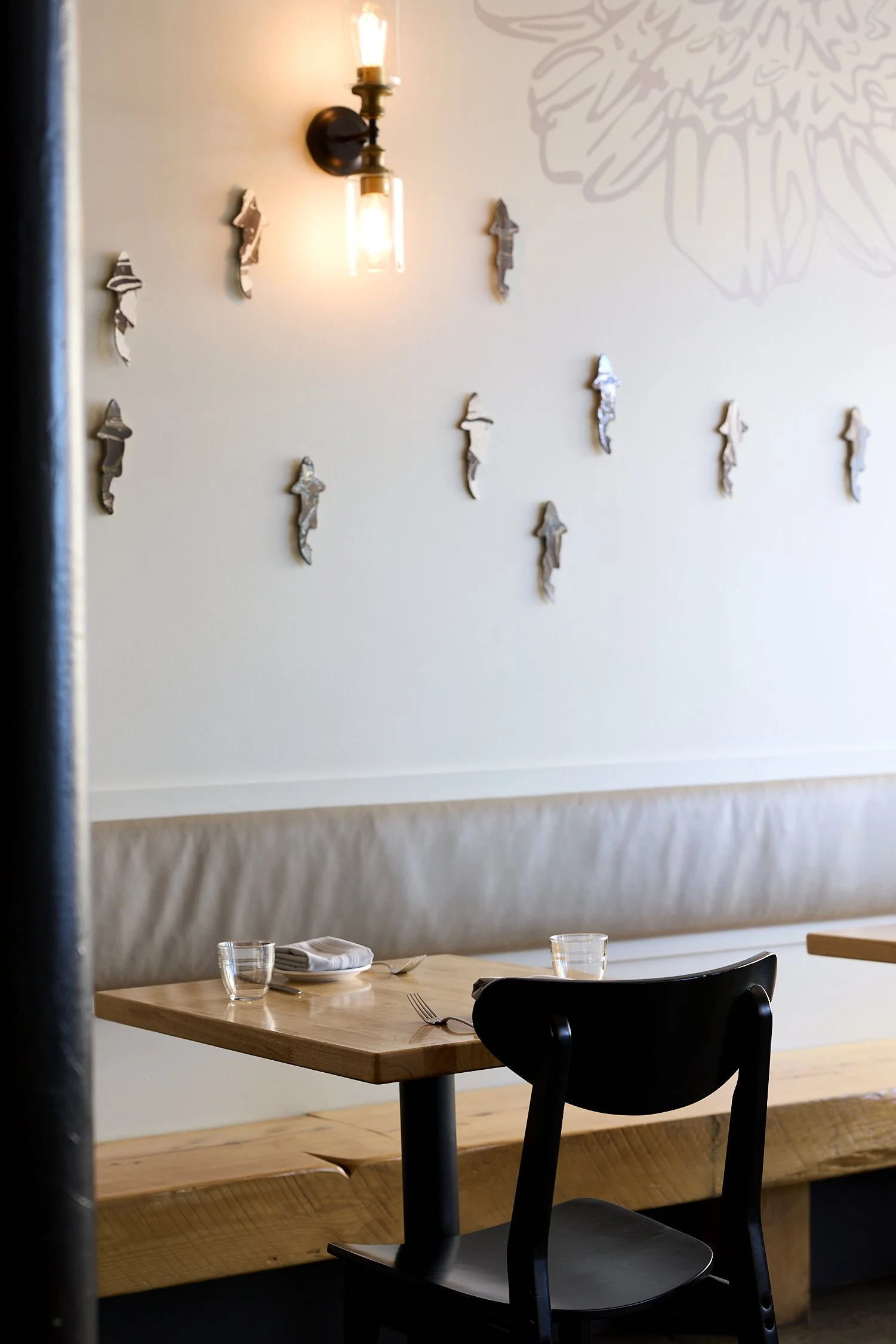 A restaurant interior with a wooden dining table set with napkins, utensils, and glasses, a black chair, and a wall decorated with a modern light fixture and scattered cutout figures.