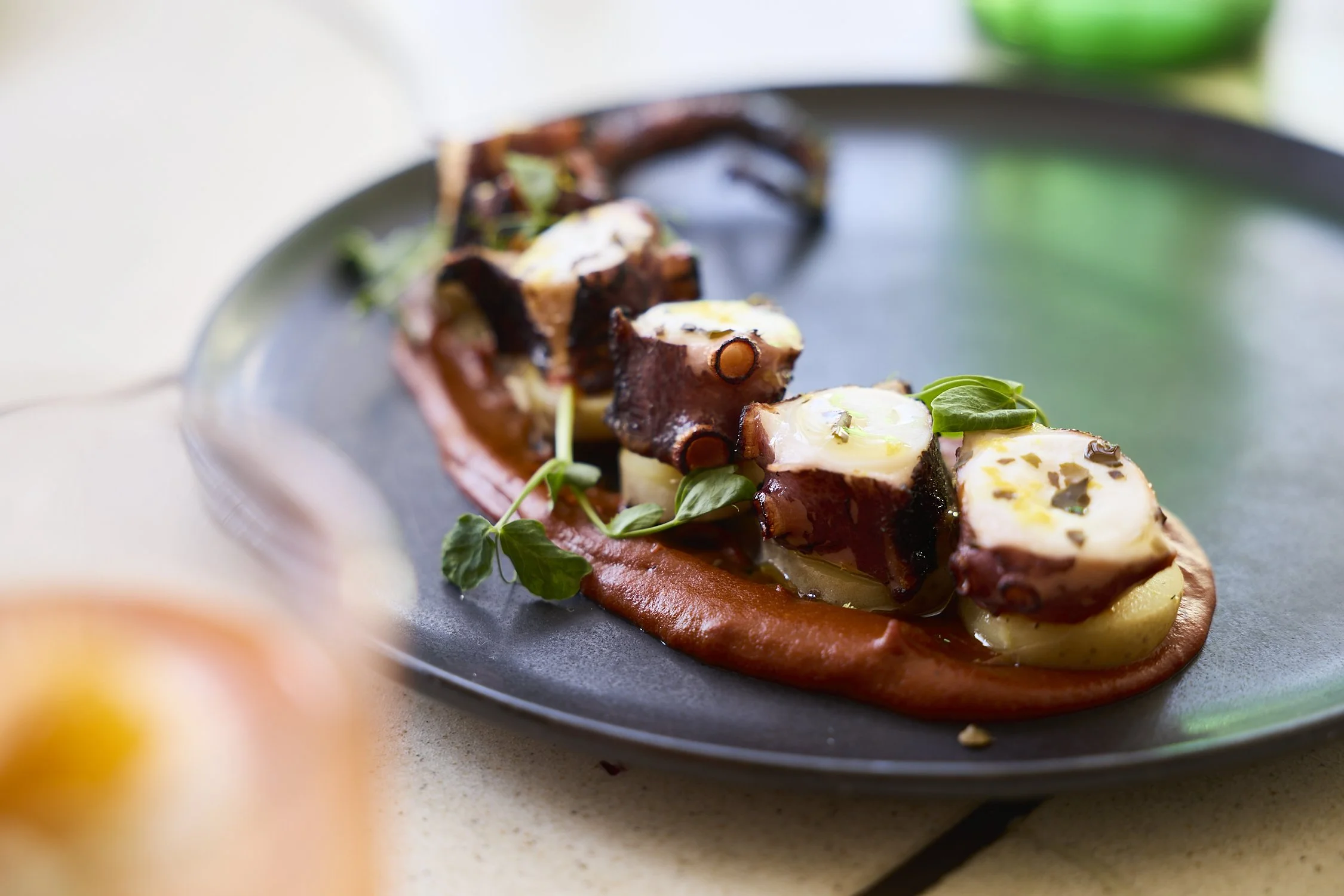 Close-up of gourmet dish with roasted octopus atop a bed of mashed potatoes, garnished with microgreens and herbs, served on a black plate.