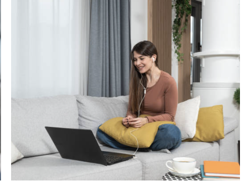 Woman sitting on a grey sofa with a yellow pillow, using a laptop and listening to music with earphones, in a cozy living room setting.