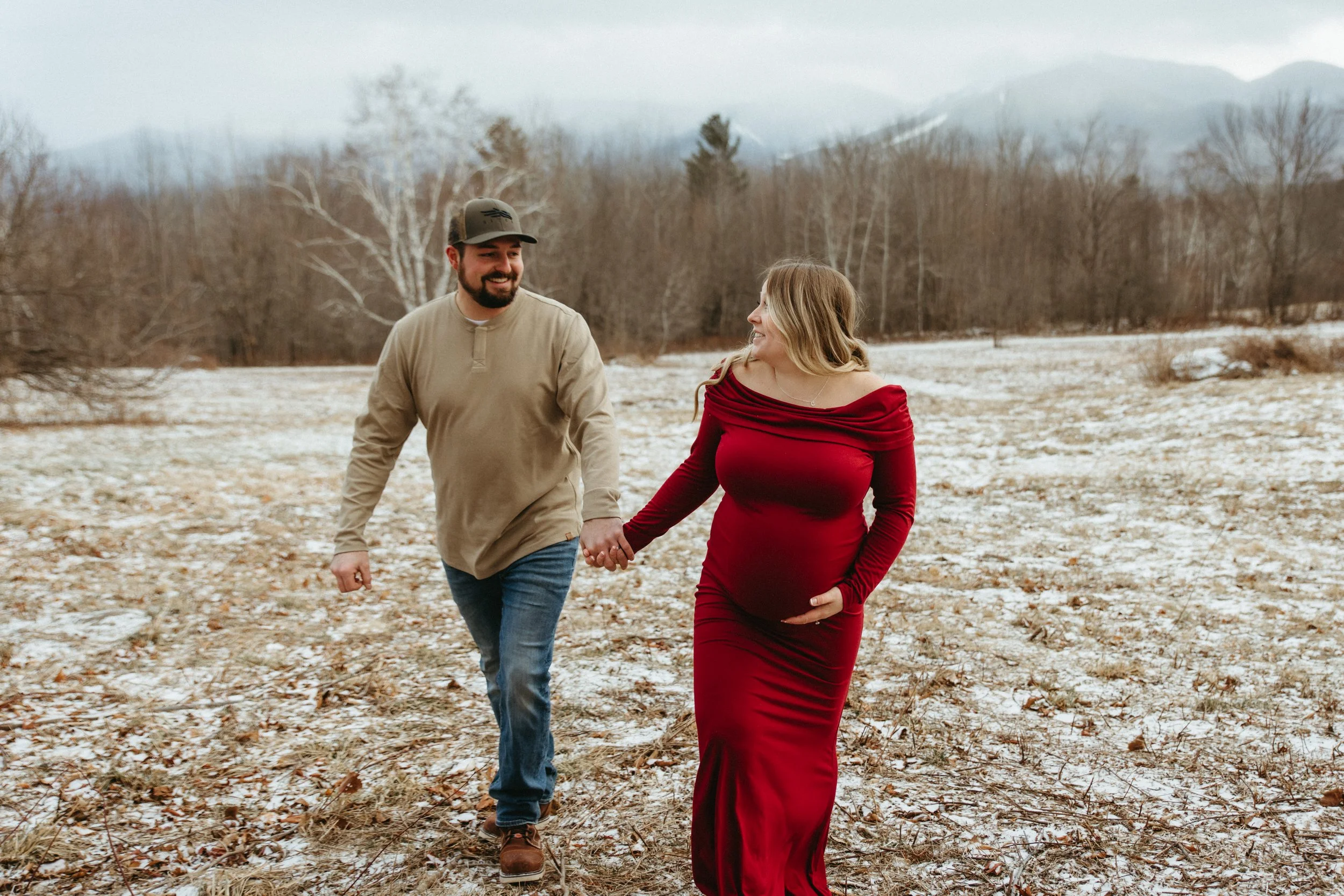 A man and woman walking hand in hand through a wintery outdoor landscape, with bare trees and distant mountains in the background. The woman is wearing a red dress and the man a beige shirt and jeans.