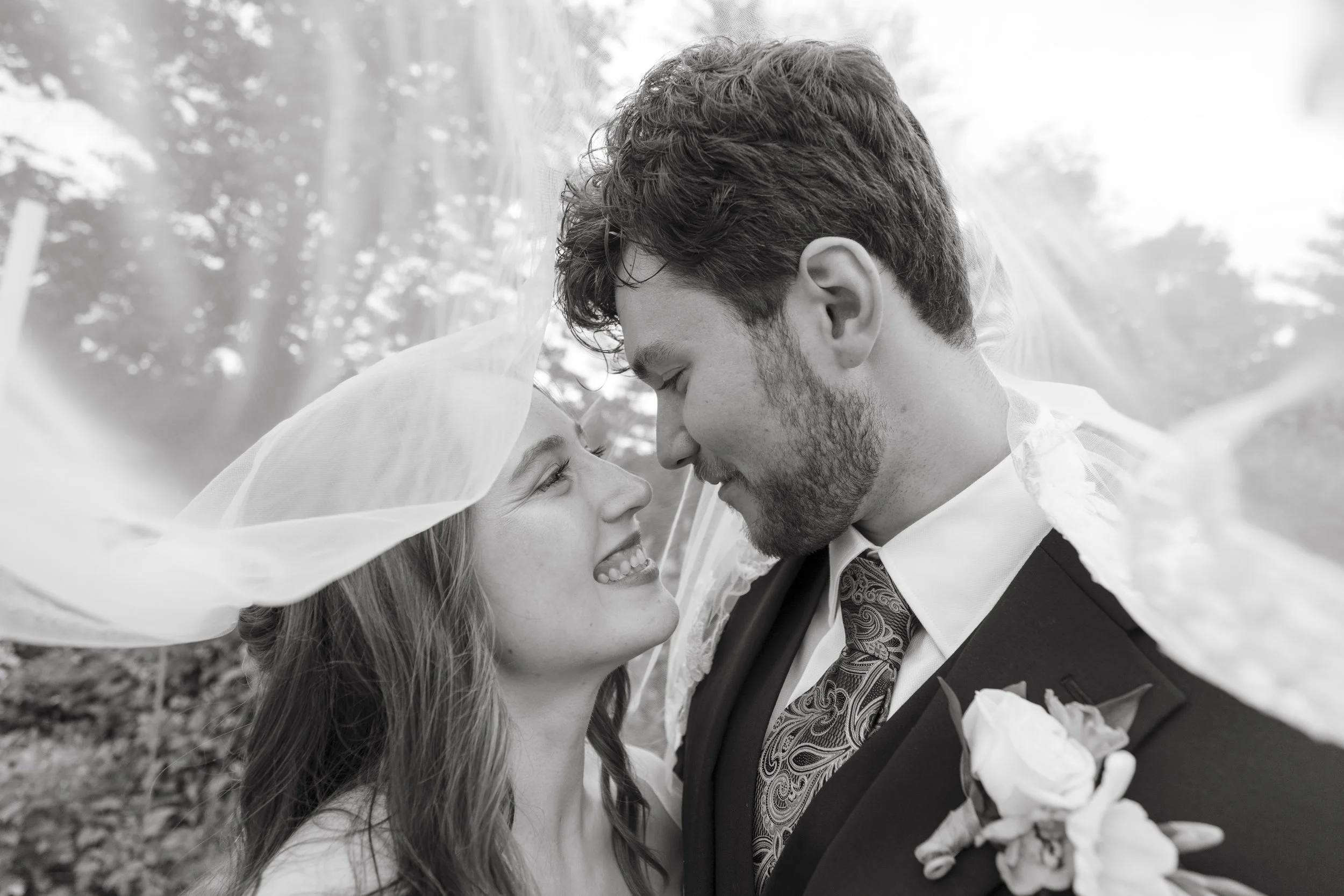 A black and white photo of a couple on their wedding day, standing outdoors closely close, gazing into each other's eyes, with trees and natural background.