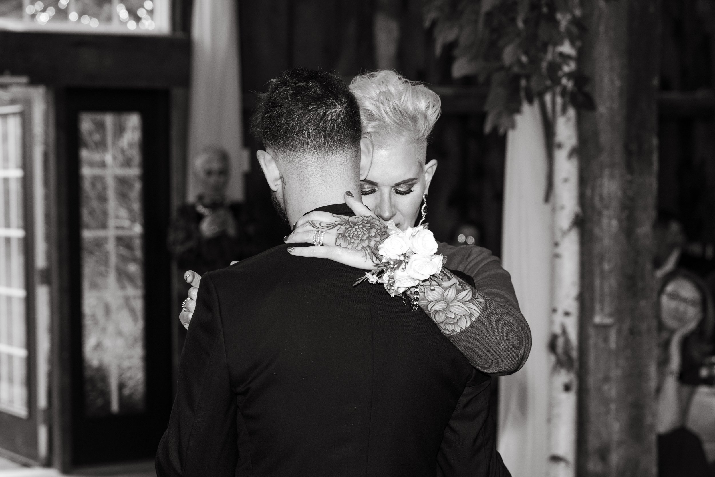 A couple is dancing closely, embracing each other; the person with short, light-colored hair has a tattooed arm and is wearing floral jewelry, while the other has short dark hair. The scene appears intimate and emotional.