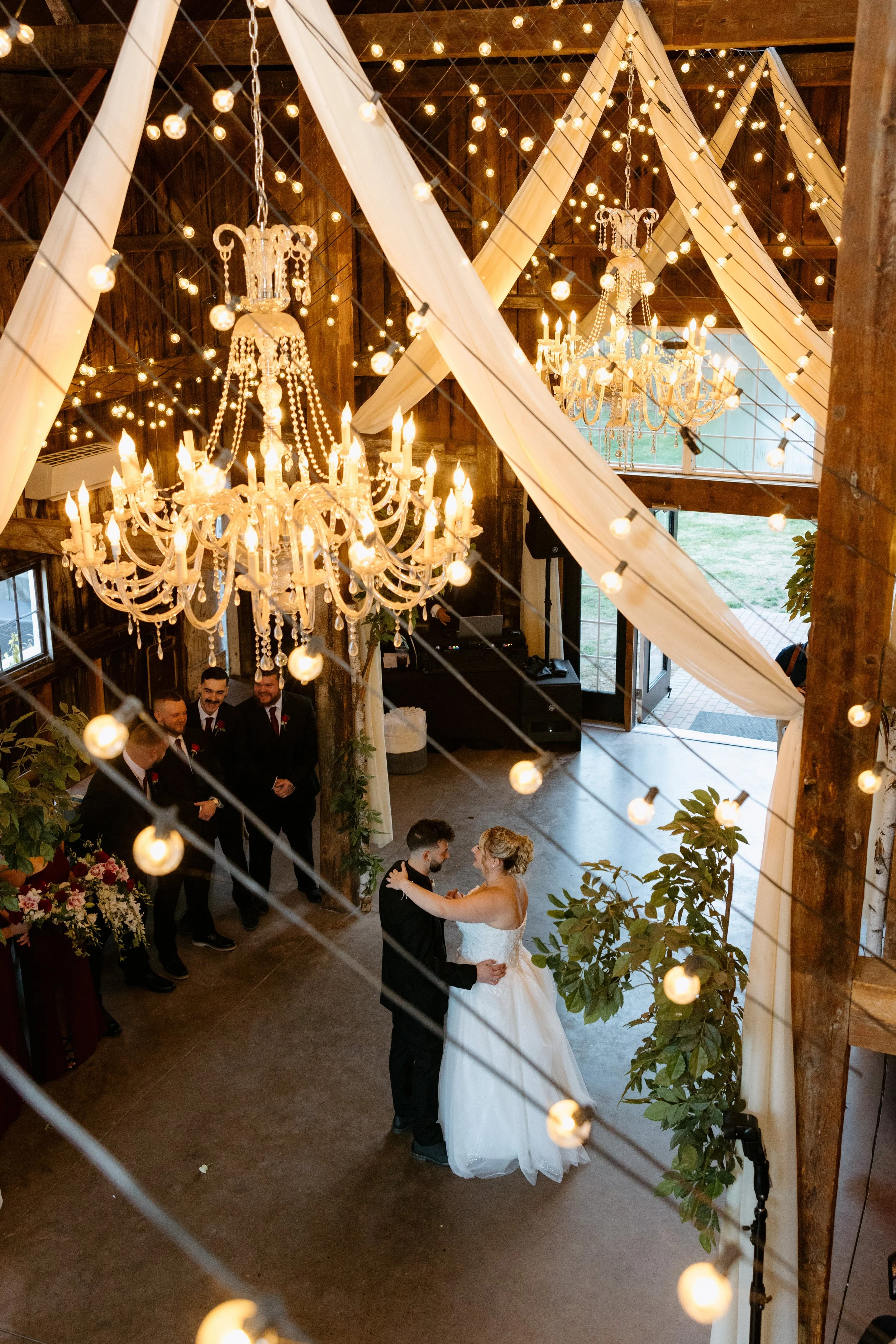 A wedding reception inside a rustic wooden barn with romantic lighting. A couple is dancing in the foreground while a group of men in suits are gathered nearby. Gorgeous chandeliers and string lights enhance the elegant ambiance.