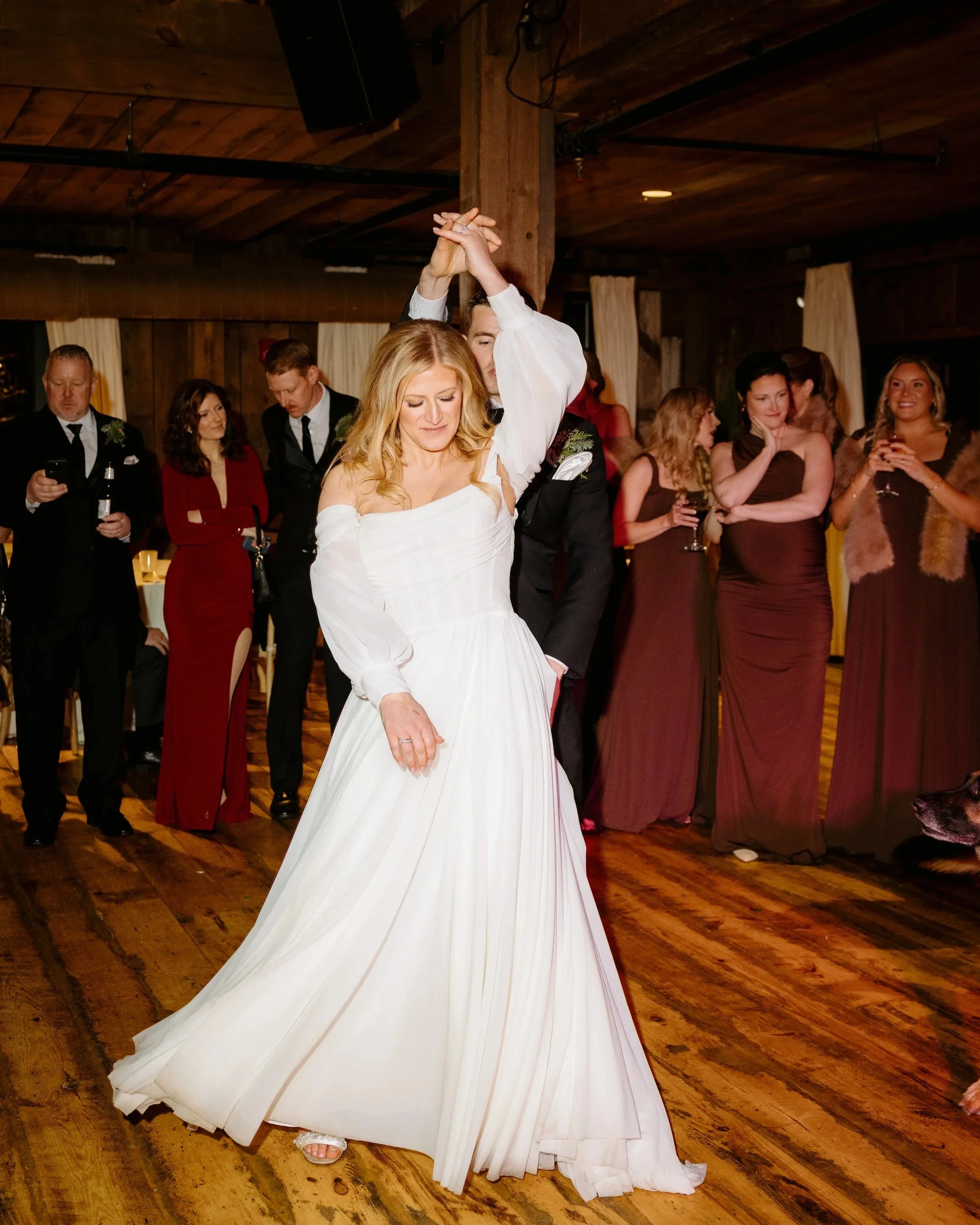 A woman in a white dress dancing with a man in a black suit at a wedding reception, with several guests watching and smiling in the background, inside a rustic venue with wooden walls and ceiling.