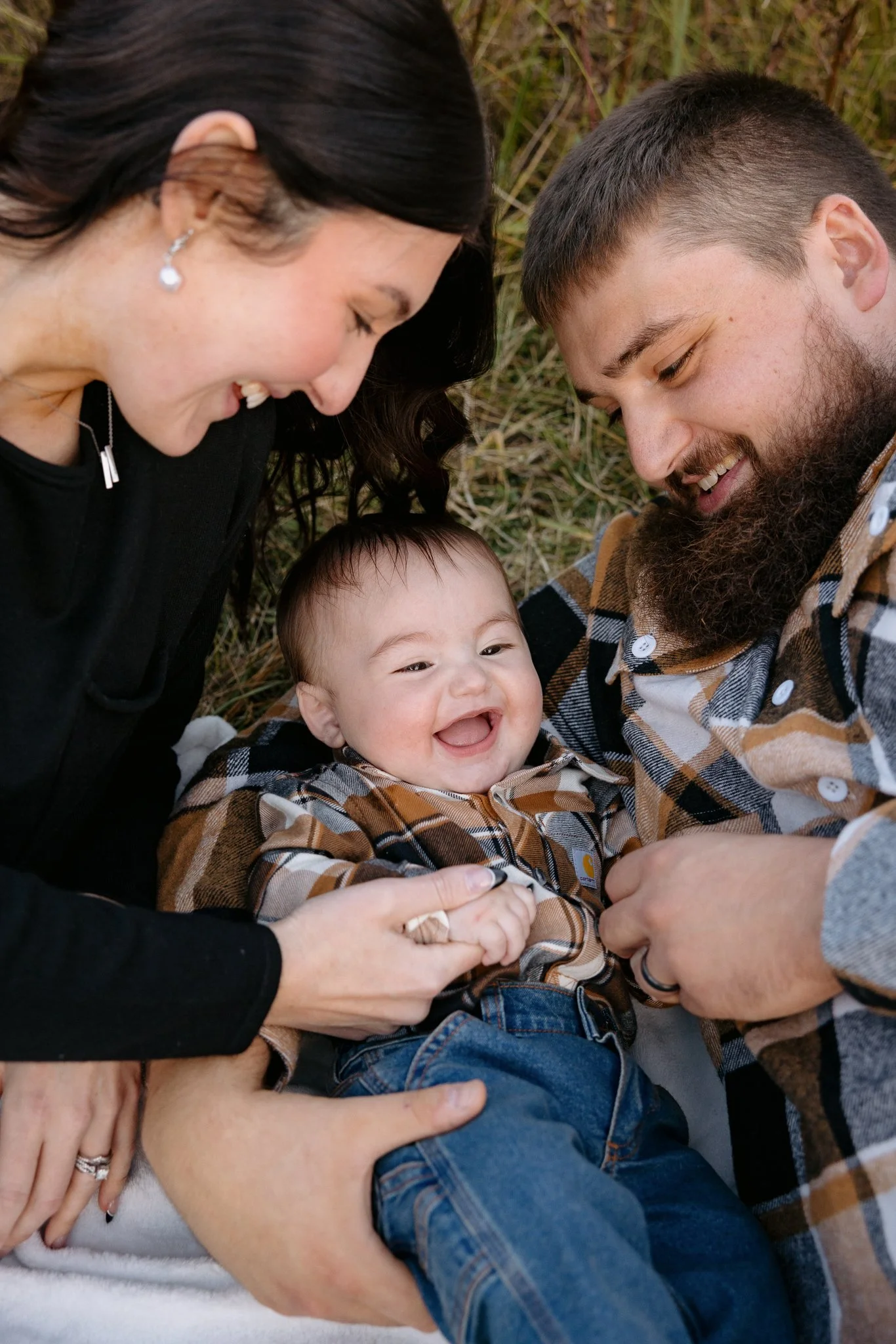 A family of four lying on grass, smiling and enjoying each other's company. The mother has dark hair and is wearing earrings, the father has a full beard, and the toddler is smiling and laughing. The family appears to be in a happy, playful moment ou