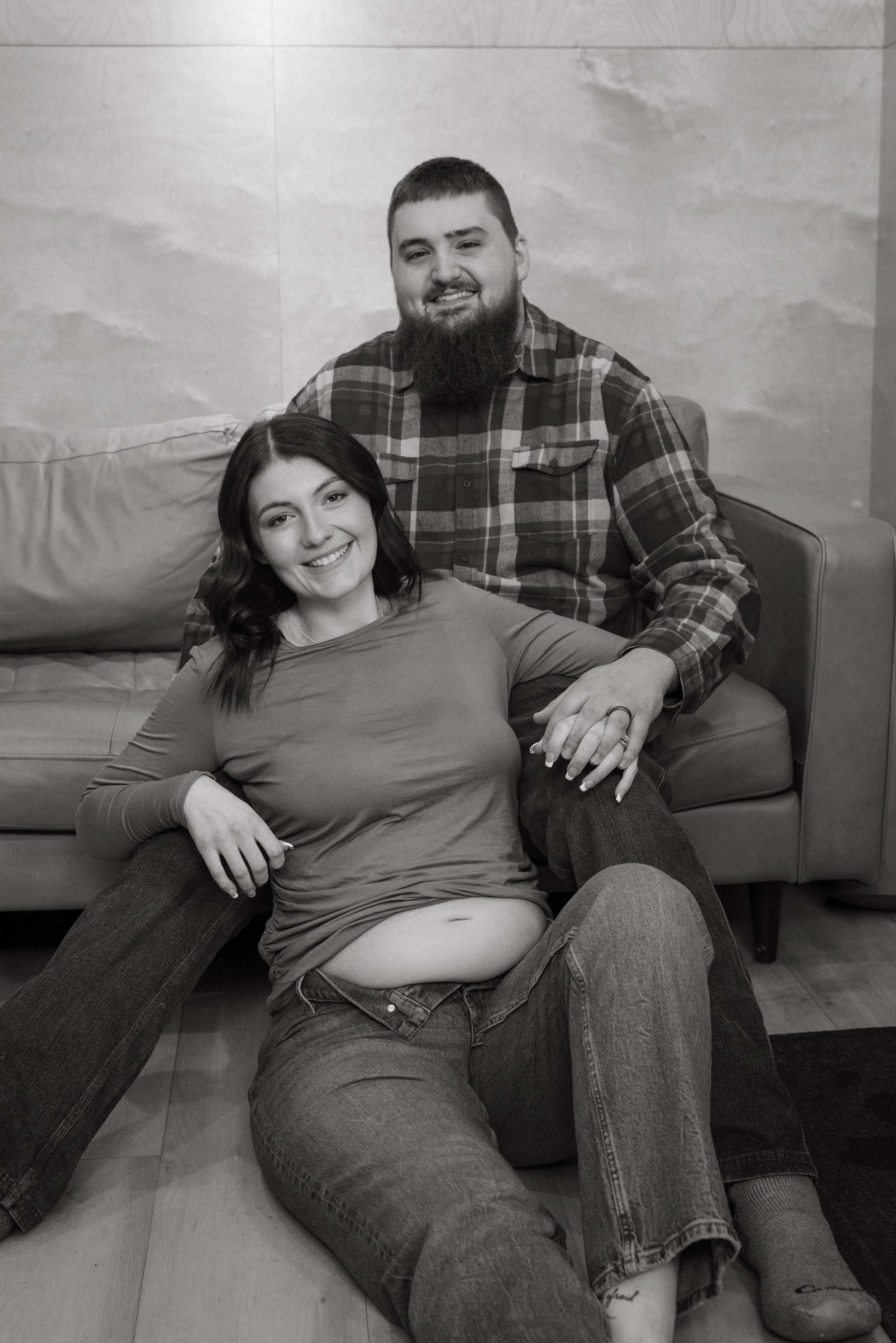 A smiling woman with dark hair and a ring on her finger is sitting on the floor with her back against a man. The man, who has a beard and is wearing a plaid shirt, is sitting on a chair behind her. They are indoors with a light-colored wall and a sof