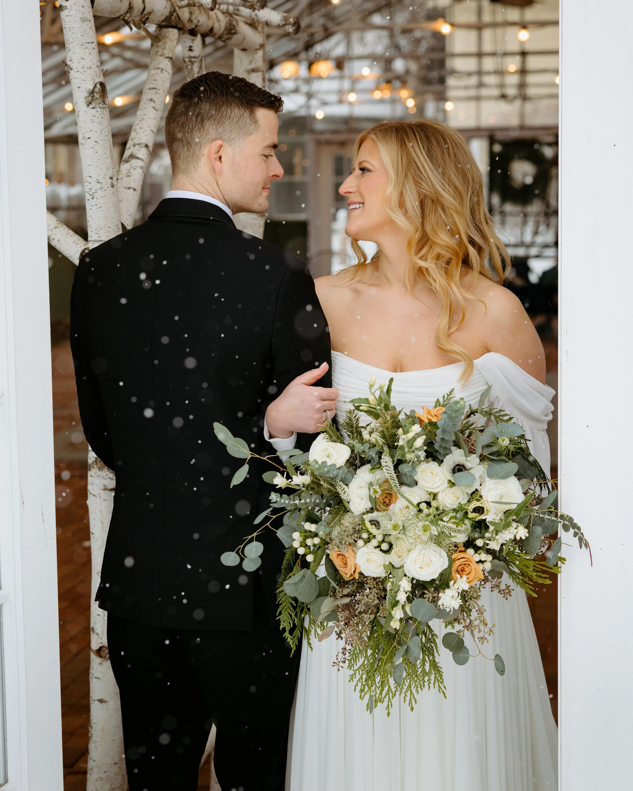 A bride and groom sharing an intimate moment in a wedding ceremony, with the bride holding a large bouquet of white and peach roses, green foliage, and other flowers, while smiling at each other inside a rustic venue decorated with string lights and 