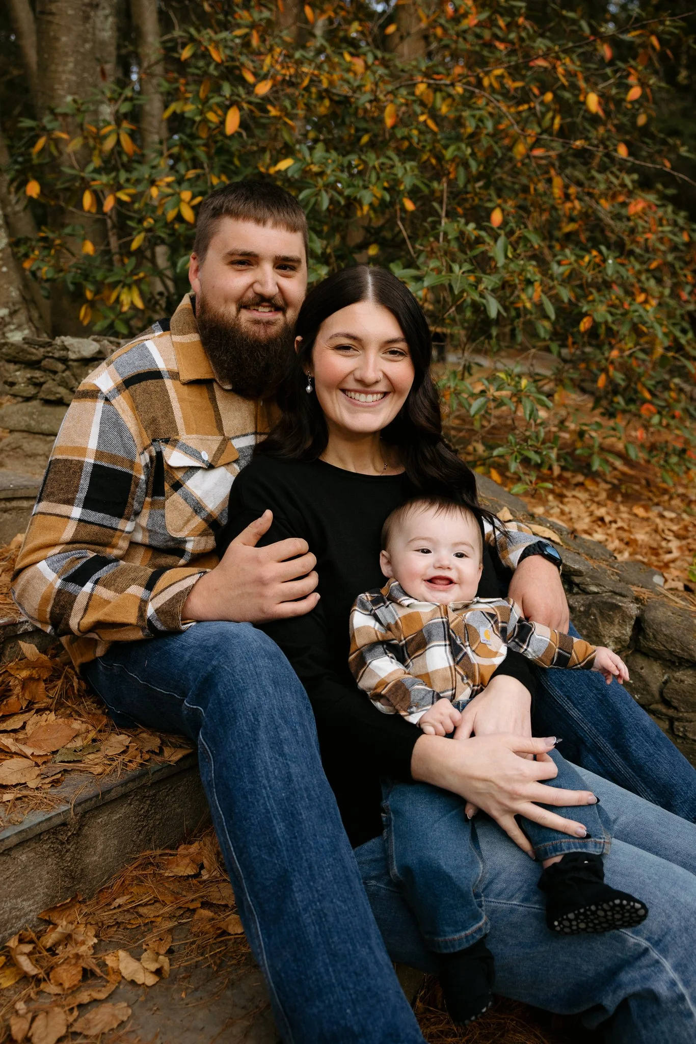 A family of three smiling and sitting outdoors during autumn, with fallen leaves on the ground and trees with orange and green leaves behind them.