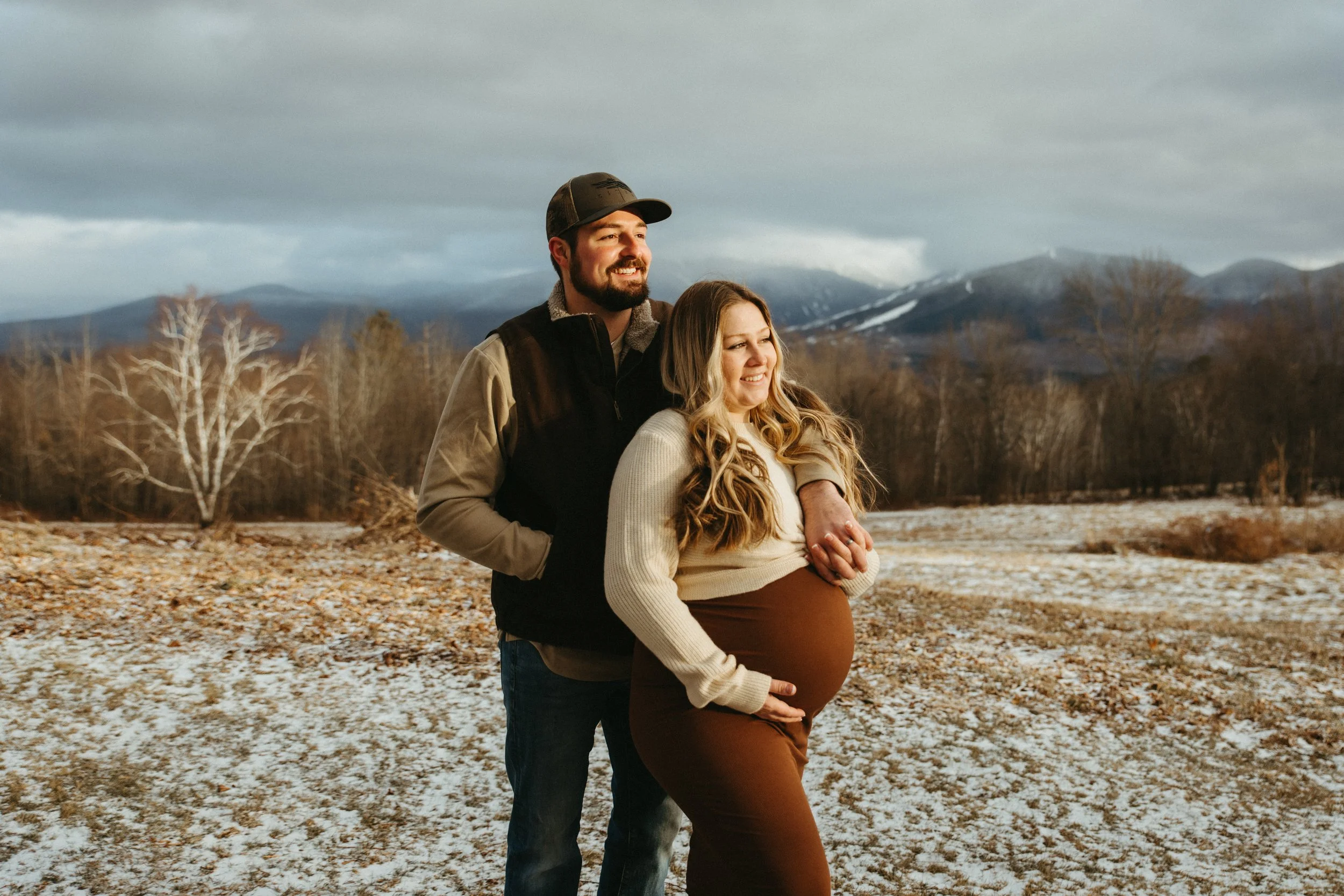 A happy couple stands together outdoors during winter, with the man embracing the pregnant woman from behind. They are smiling and looking into the distance with snow-covered mountains and leafless trees in the background.