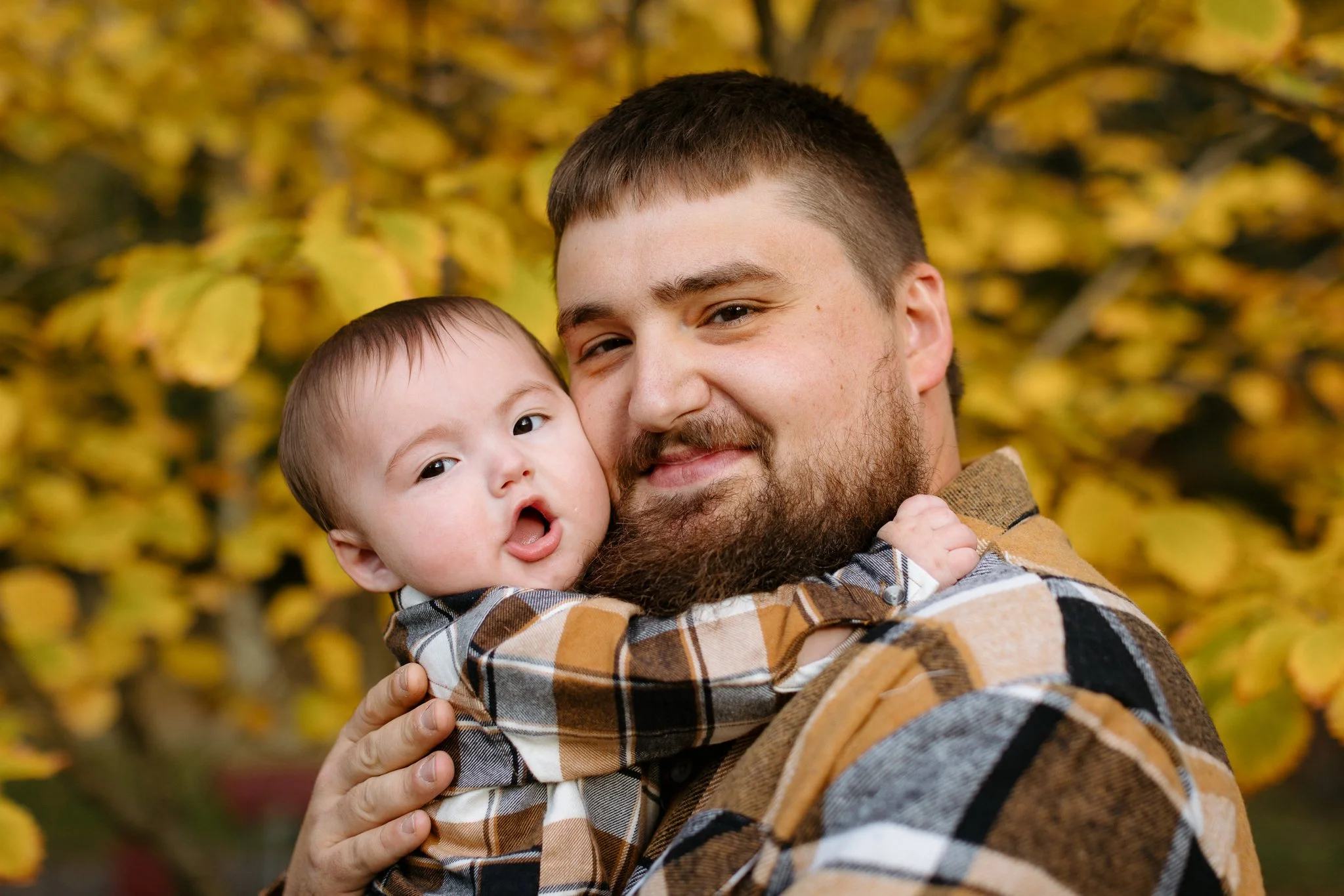 A man with a beard holding a young child close, both smiling, with yellow fall leaves in the background.