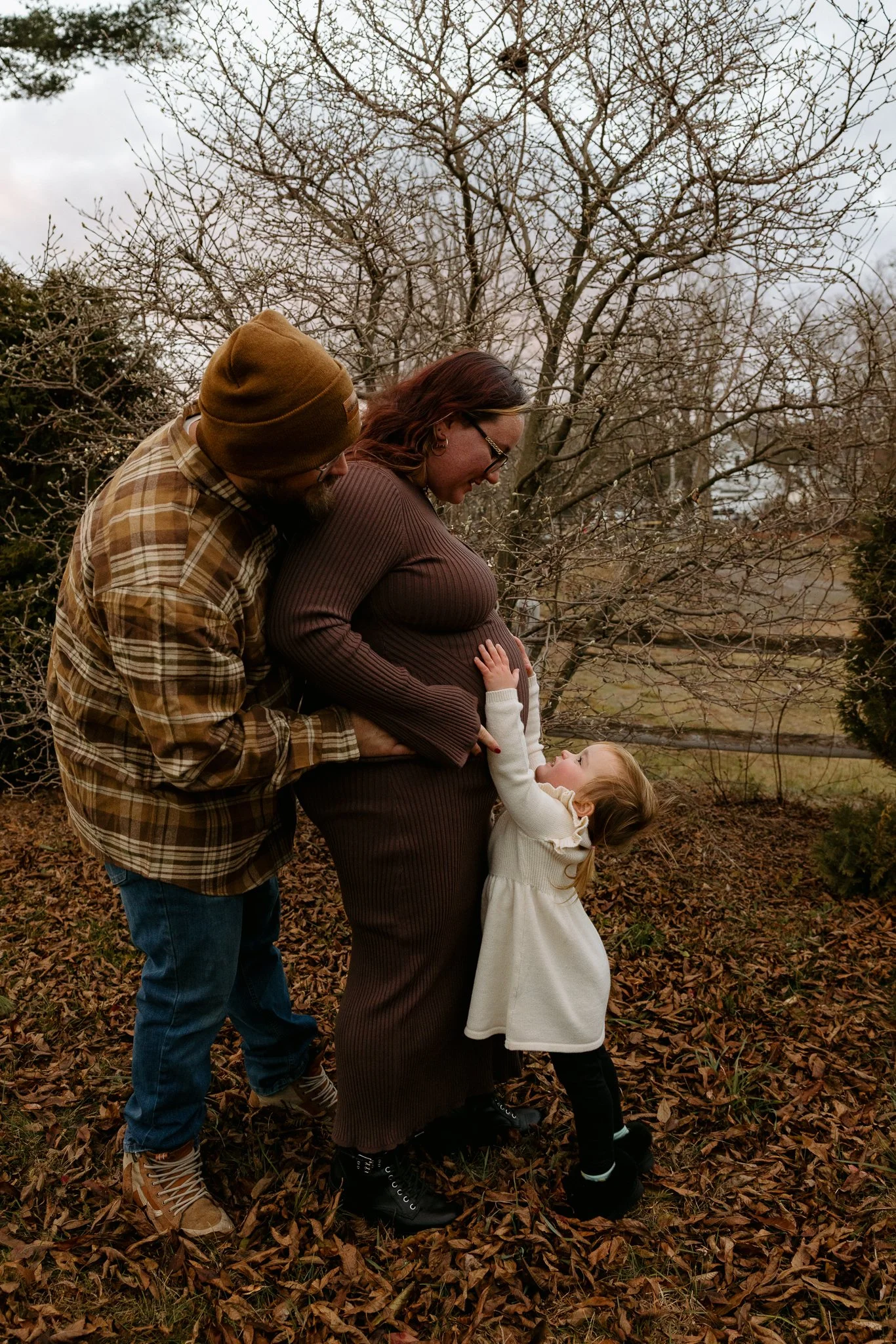 A family of four standing outdoors in a wooded area during fall, with brown leaves on the ground and a leafless tree in the background. The mother, with red hair and glasses, is pregnant and smiling, while the young daughter, wearing a white dress, i
