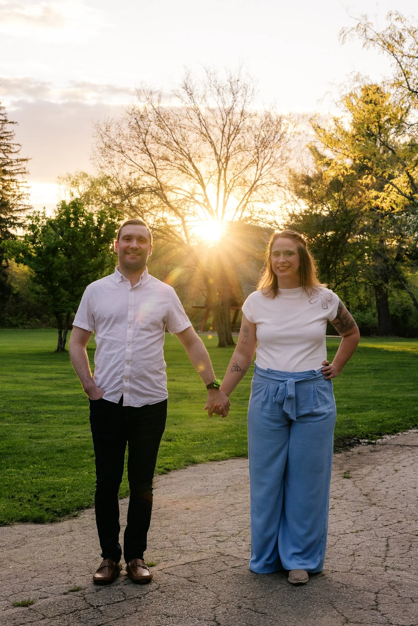 A man and woman walking hand in hand in a park during sunset, with trees in the background and a cracked paved path beneath them.