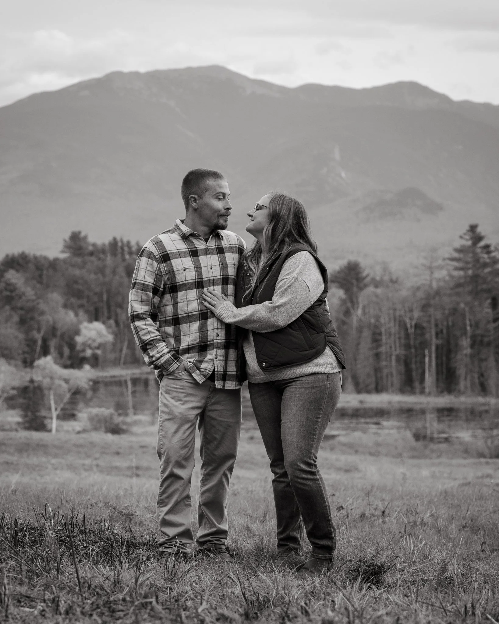 A black-and-white photo of a couple standing outdoors in a grassy area, embracing each other, with a mountain range in the background.