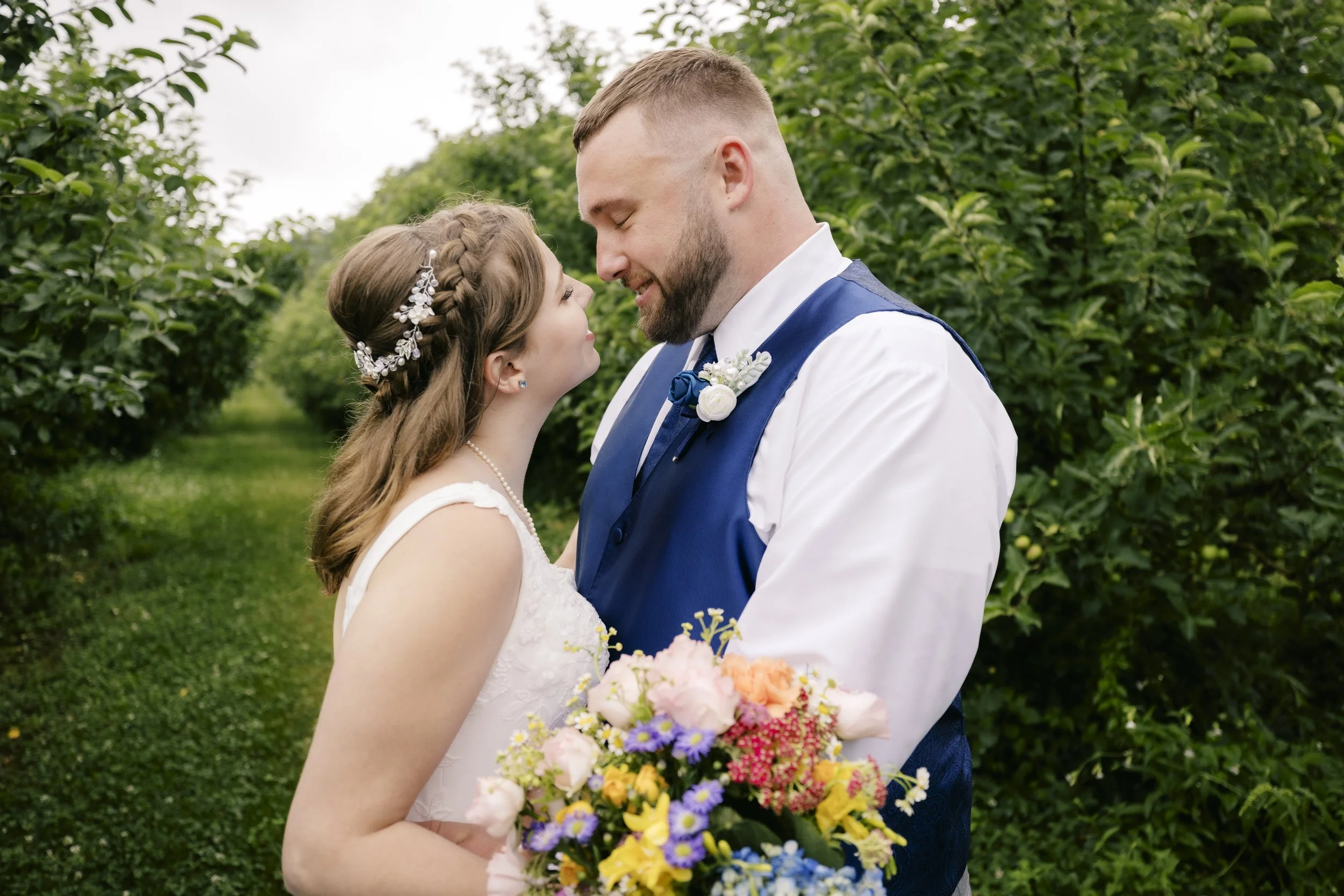 A bride and groom standing close together outdoors, smiling at each other, with a green hedge background. The bride is holding a colorful bouquet of flowers, and the groom is wearing a blue vest and tie with a white shirt.