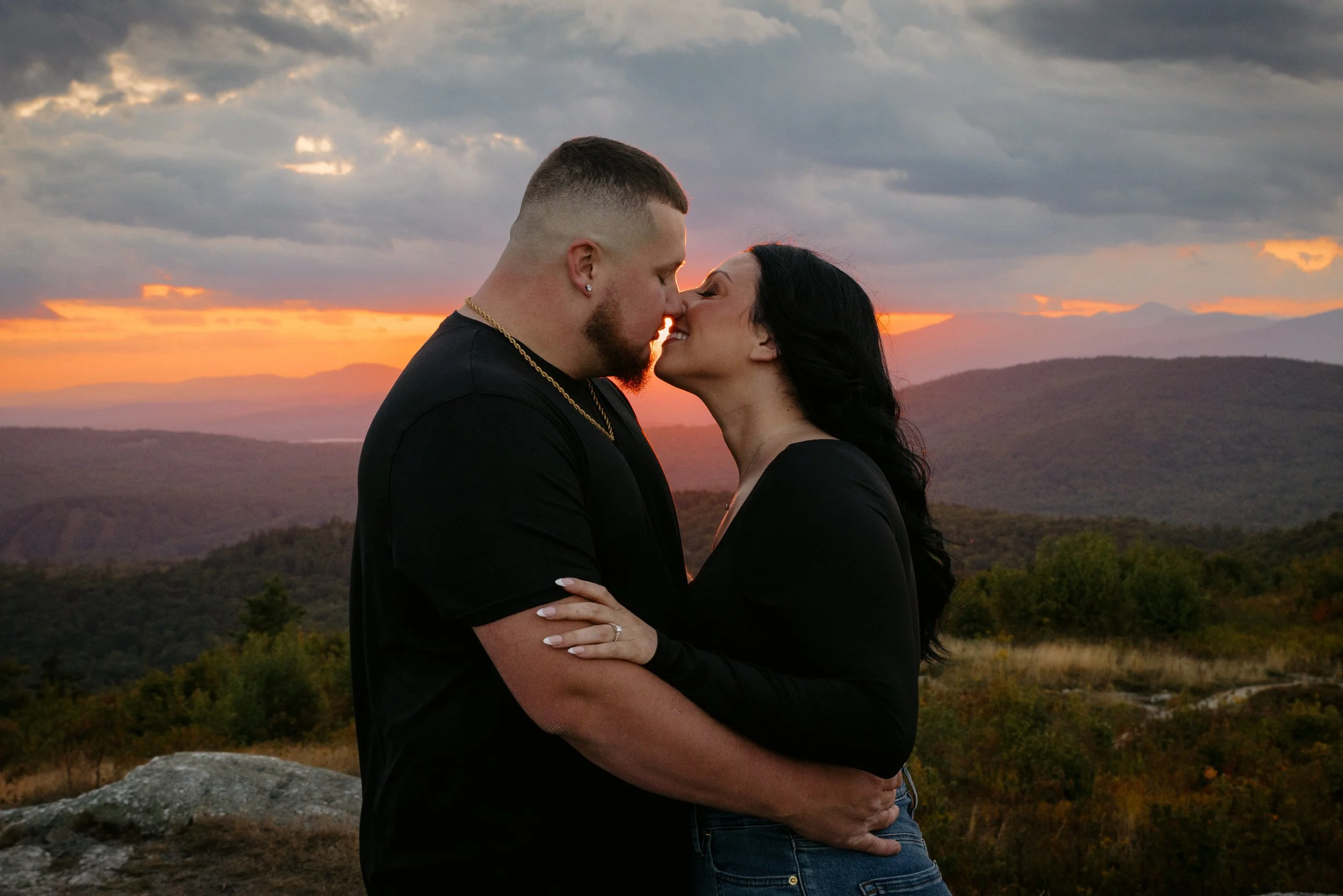 A man and woman are embracing each other outdoors during sunset, with mountains and a cloudy sky in the background.