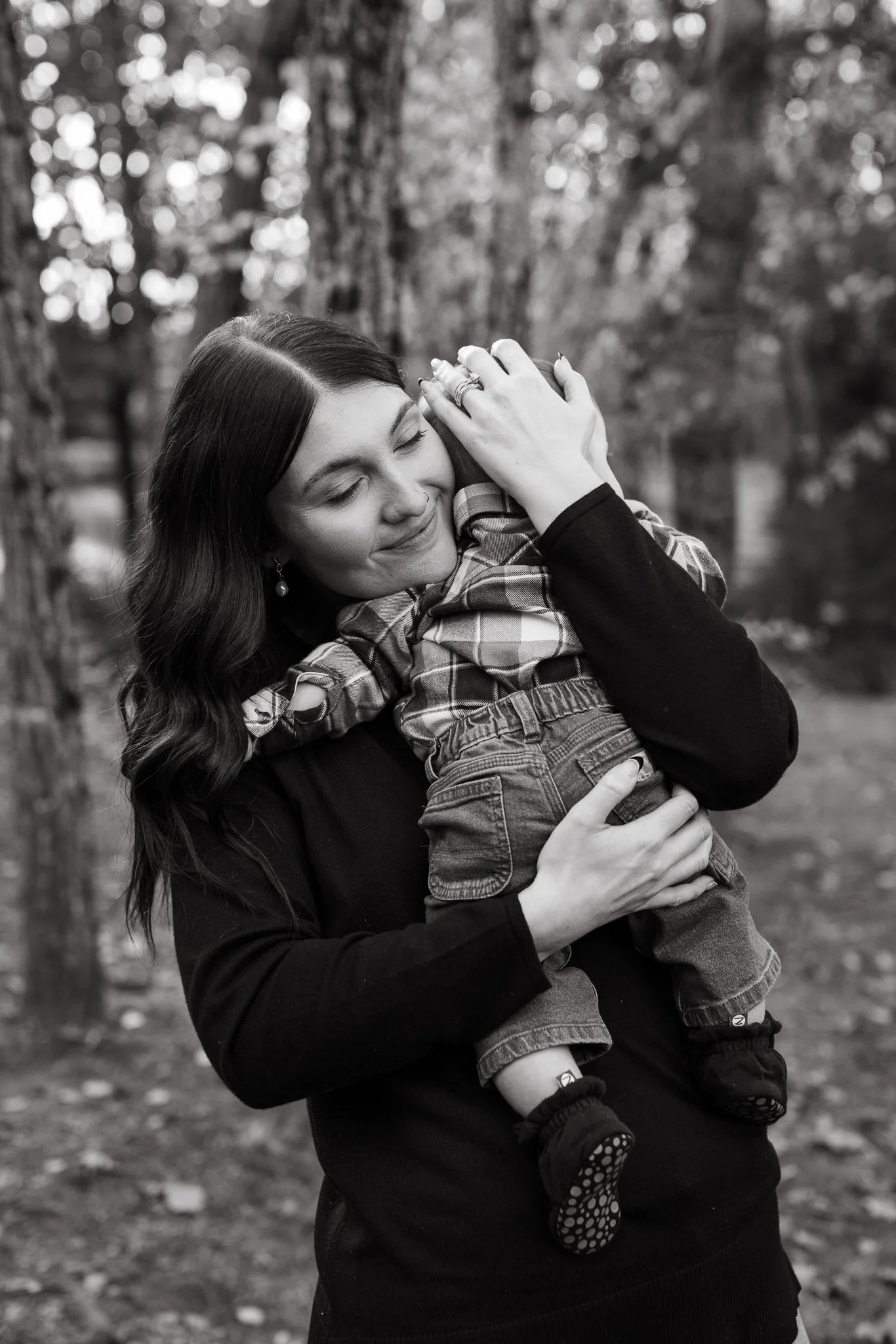 A woman with wavy hair, wearing earrings and a dark top, is smiling and hugging a young girl with long hair, wearing a plaid shirt, jeans, and patterned socks. They are outdoors in a wooded area.