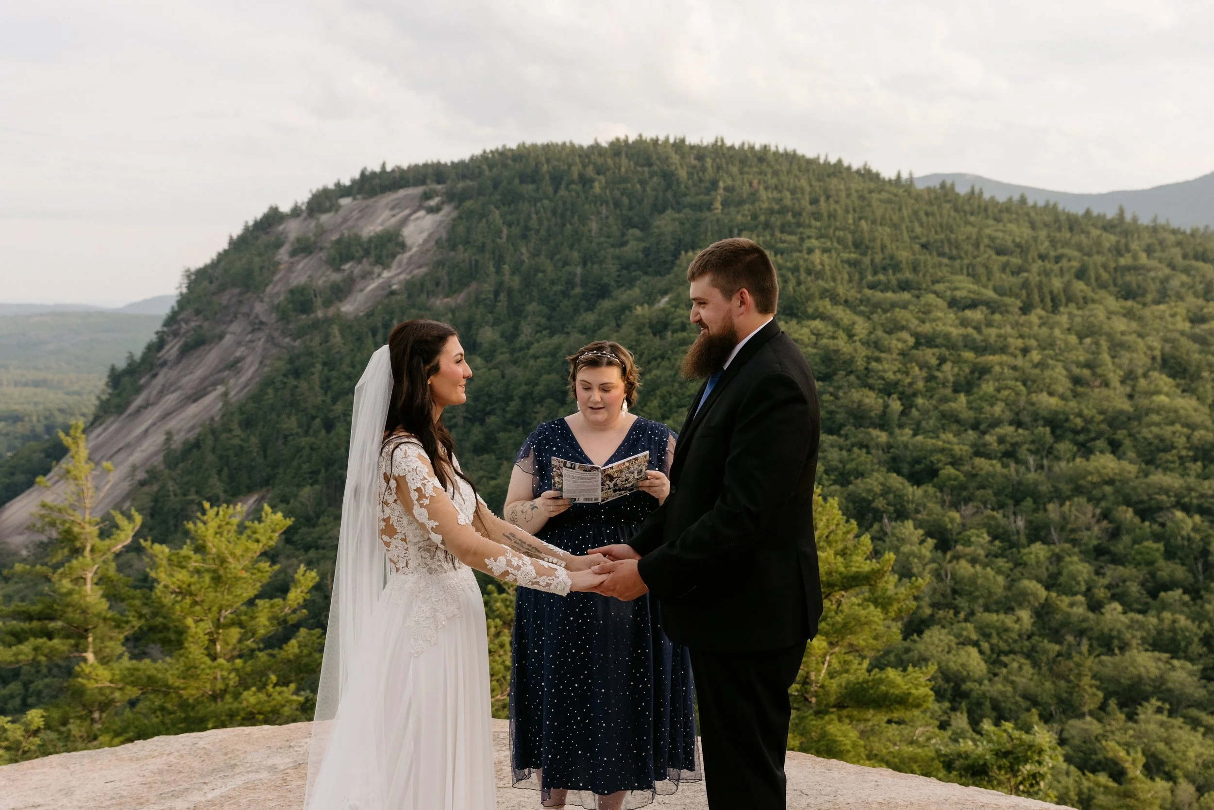A wedding ceremony taking place outdoors with a bride and groom holding hands, an officiant reading from a booklet, and a scenic mountain backdrop.